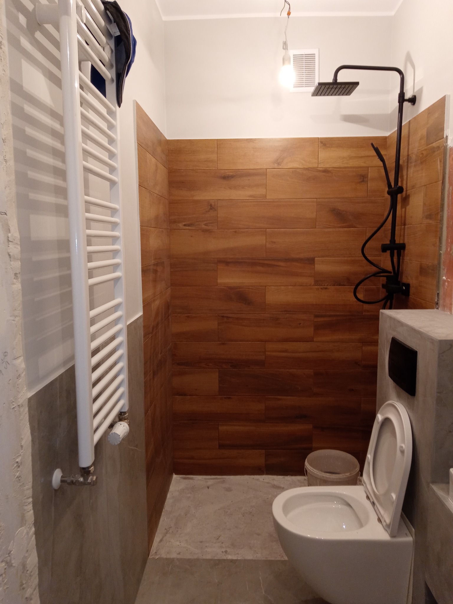 Niedokończona łazienka z drewnopodobnymi płytkami, białą toaletą wiszącą z otwartą klapą, czarną deszczownicą i białym grzejnikiem drabinkowym, widoczne niedociągnięcia w wykończeniu.