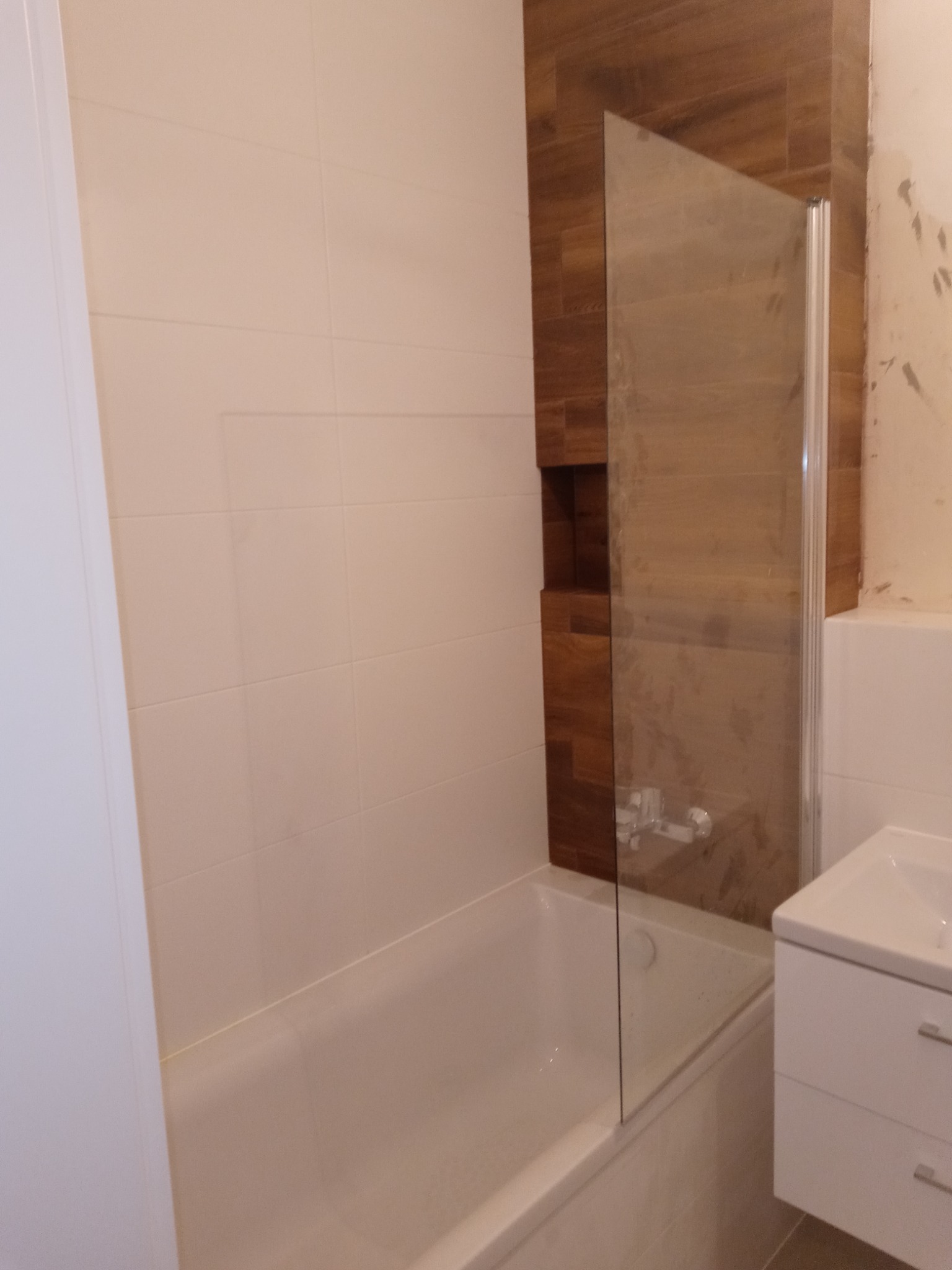 Biała wanna z zamontowanym szklanym parawanem, obok umywalka z szafką, ściany wyłożone jasnymi płytkami, fragment ściany z drewnopodobnymi płytkami z wnęką.