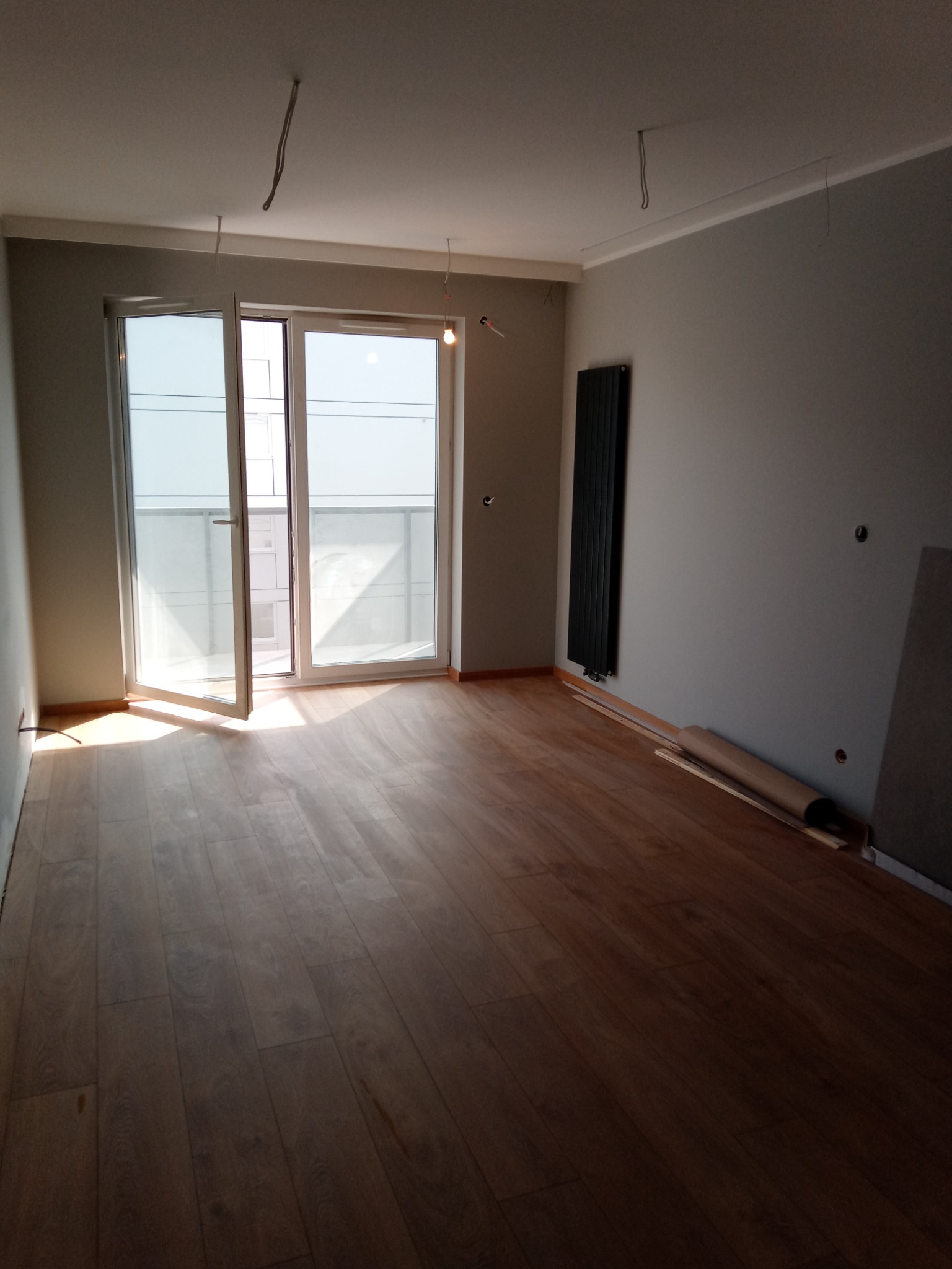 Pokój po położeniu paneli podłogowych, widoczne odstające kable ze ściany i sufitu, grzejnik panelowy na ścianie, rozłożony karton zabezpieczający podłogę.