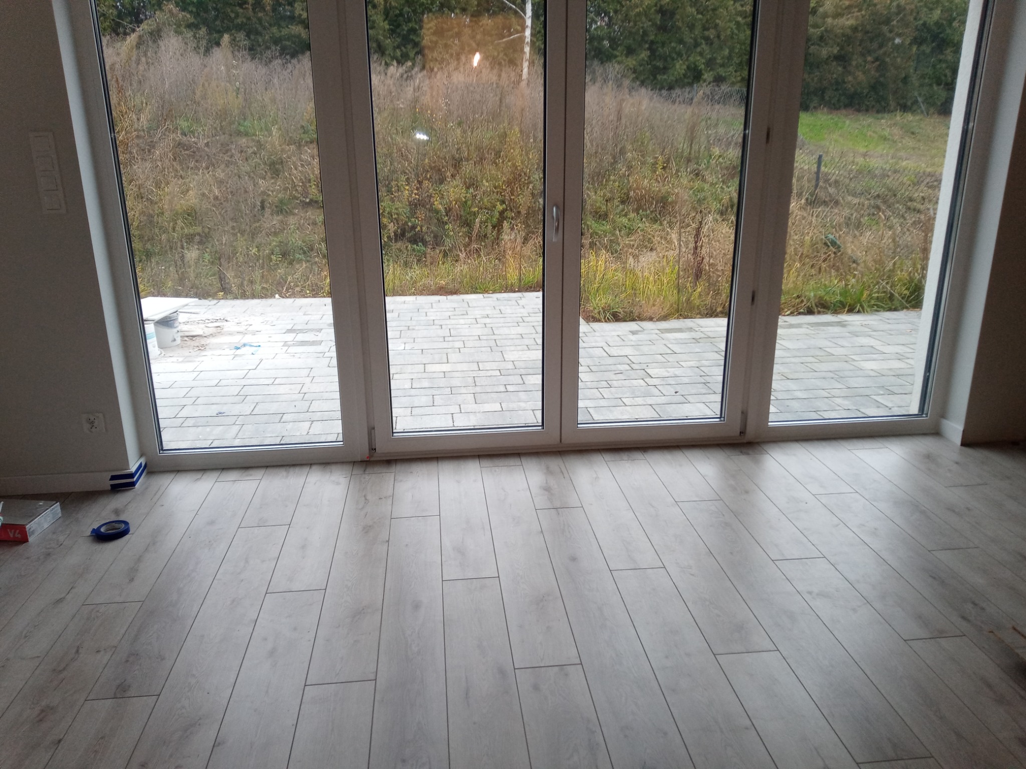Jasne panele podłogowe ułożone w nowym domu, widoczne przez duże okno tarasowe z widokiem na ogród z kostką brukową, niebieska taśma malarska i opakowanie paneli leżące na podłodze.