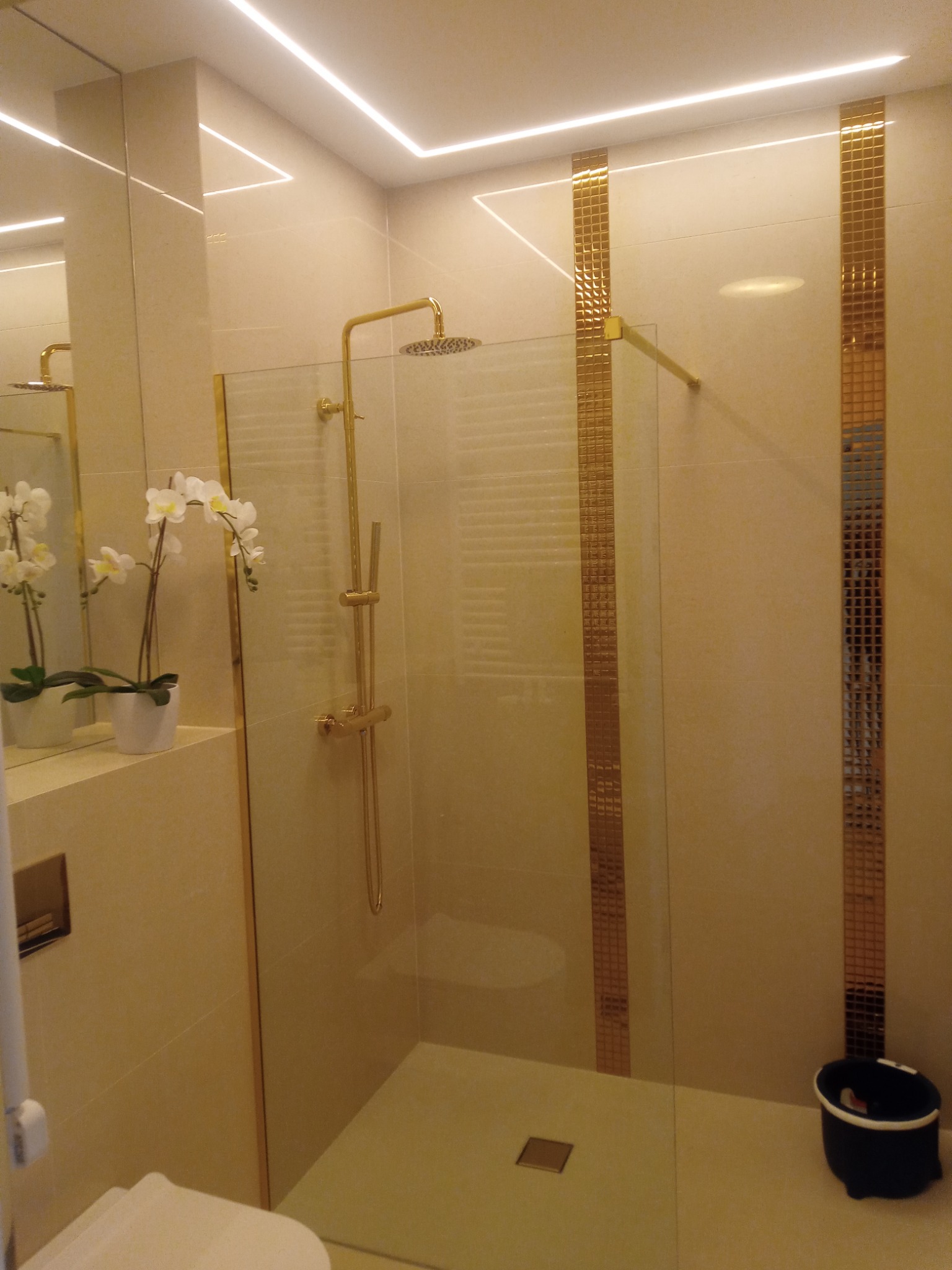 Nowoczesna kabina prysznicowa z złotymi akcentami, szklaną ścianą i liniowym oświetleniem LED w górnej części, uzupełniona dekoracyjną orchideą na umywalce.