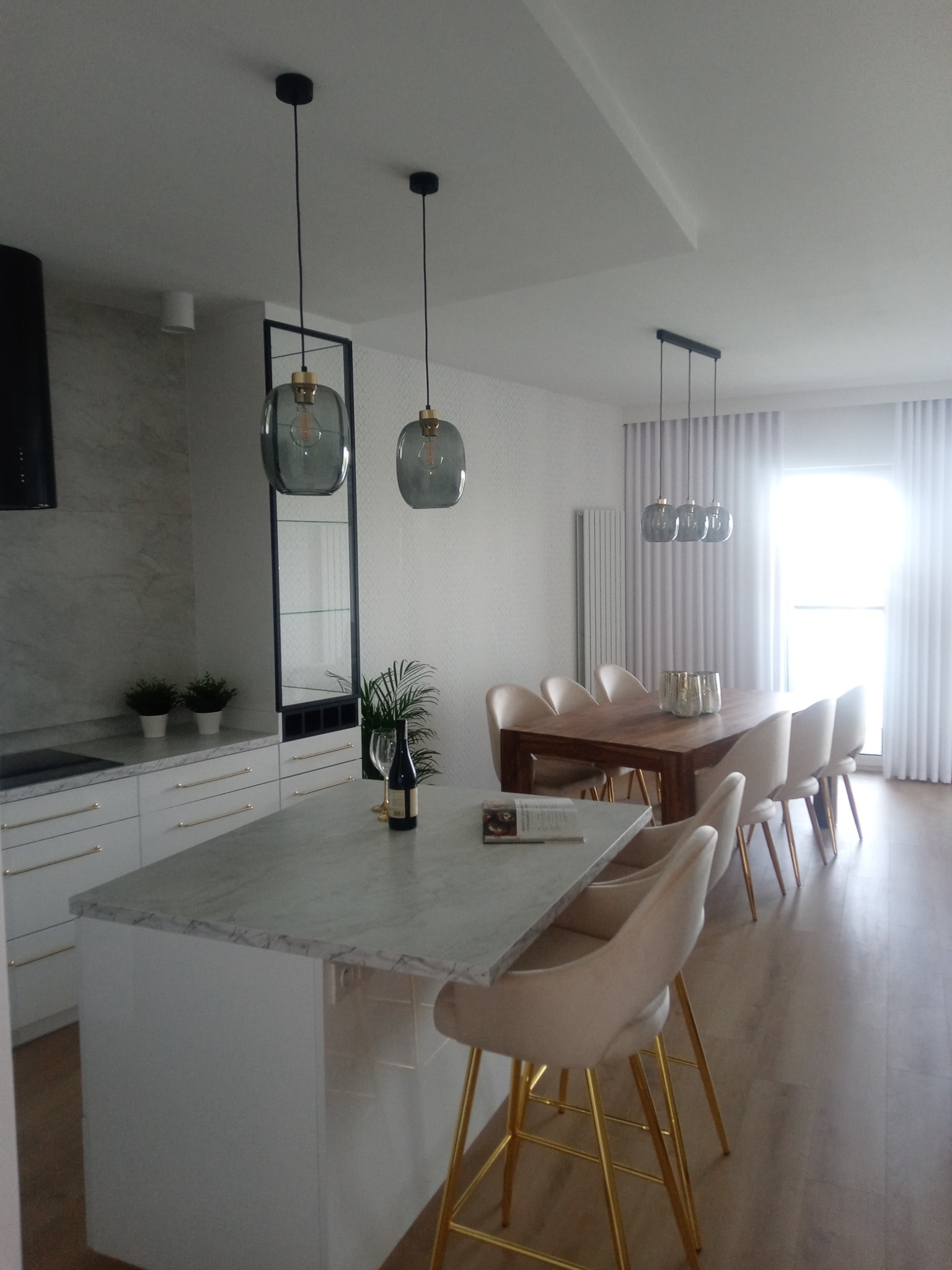 Jasne wnętrze: kuchnia z wyspą i marmurowym blatem, szklane lampy wiszące, stół z krzesłami w stylu glamour, subtelna tapeta w tle. Butelka wina i kieliszek na wyspie.