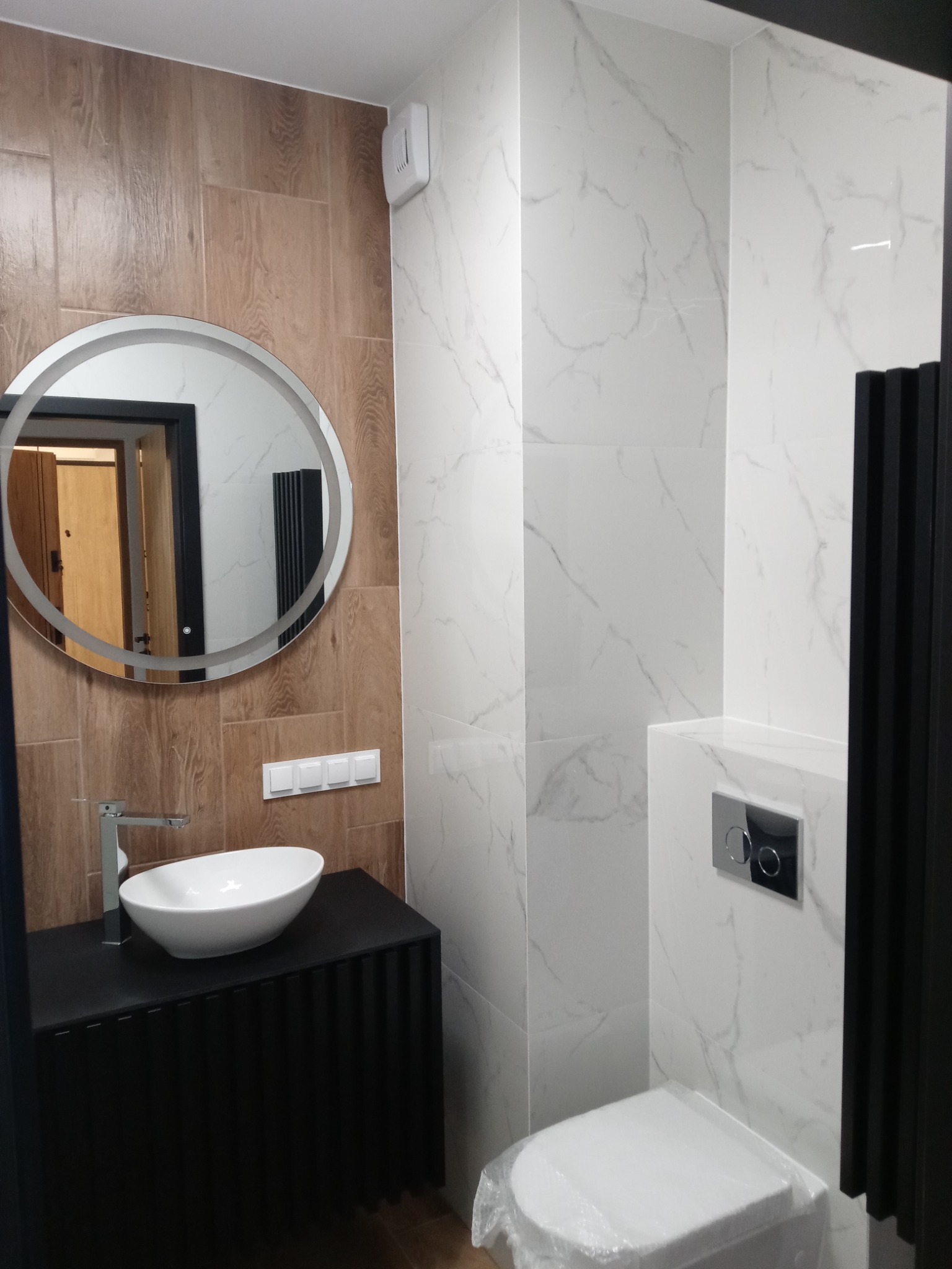 Nowoczesna łazienka z okrągłym lustrem podświetlanym LED, białą umywalką nablatową na czarnej szafce z ryflowanym frontem, drewnopodobnymi i marmurowymi płytkami oraz wiszącą miską WC z chromowanym...