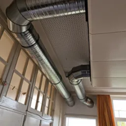 Dwa spiralne kanały wentylacyjne z ocynkowanej blachy, zamontowane pod perforowanym sufitem w jasnym pomieszczeniu z oknami i beżową zasłoną, widoczna drabina aluminiowa.