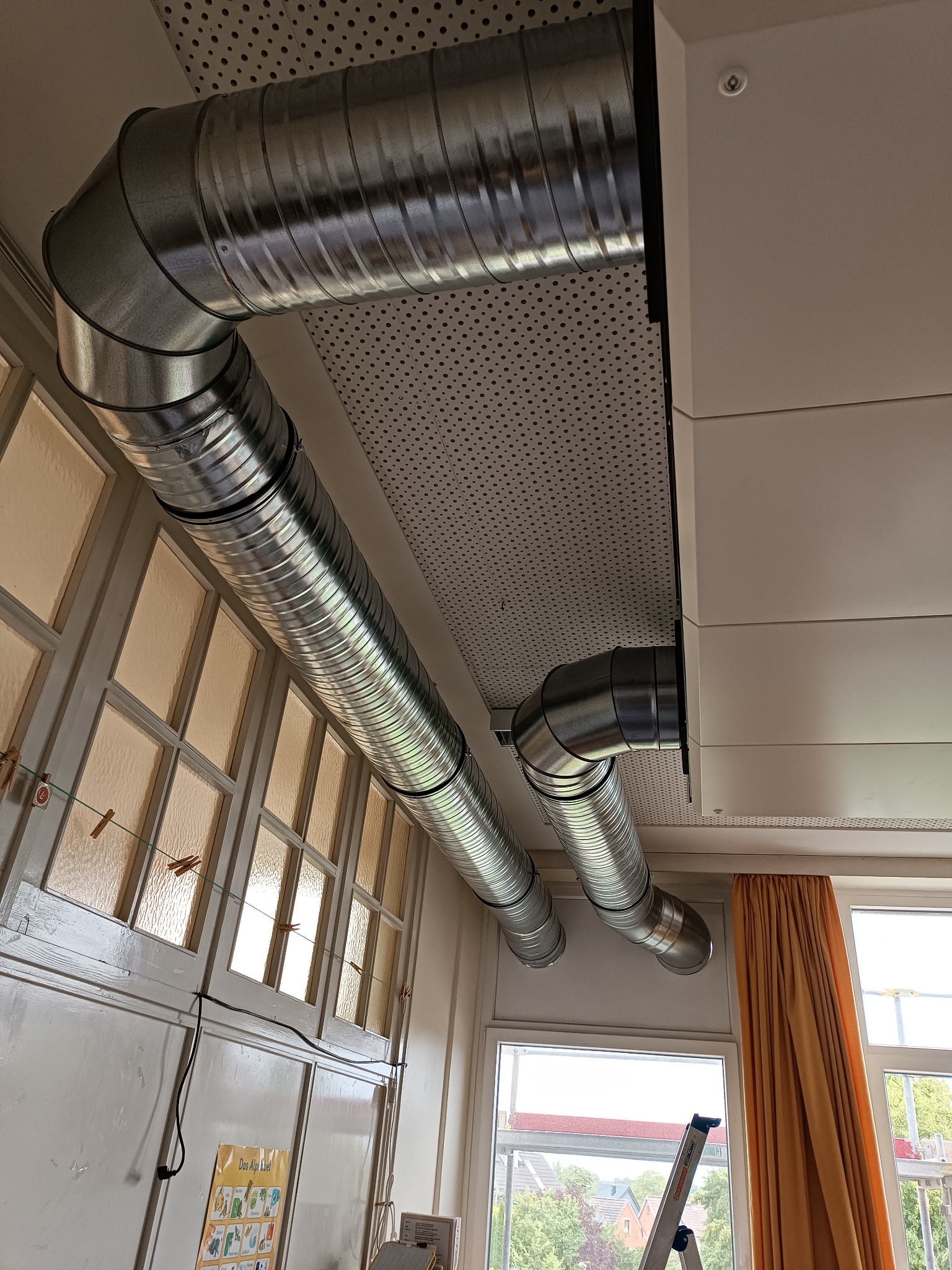 Dwa spiralne kanały wentylacyjne z ocynkowanej blachy, zamontowane pod perforowanym sufitem w jasnym pomieszczeniu z oknami i beżową zasłoną, widoczna drabina aluminiowa.