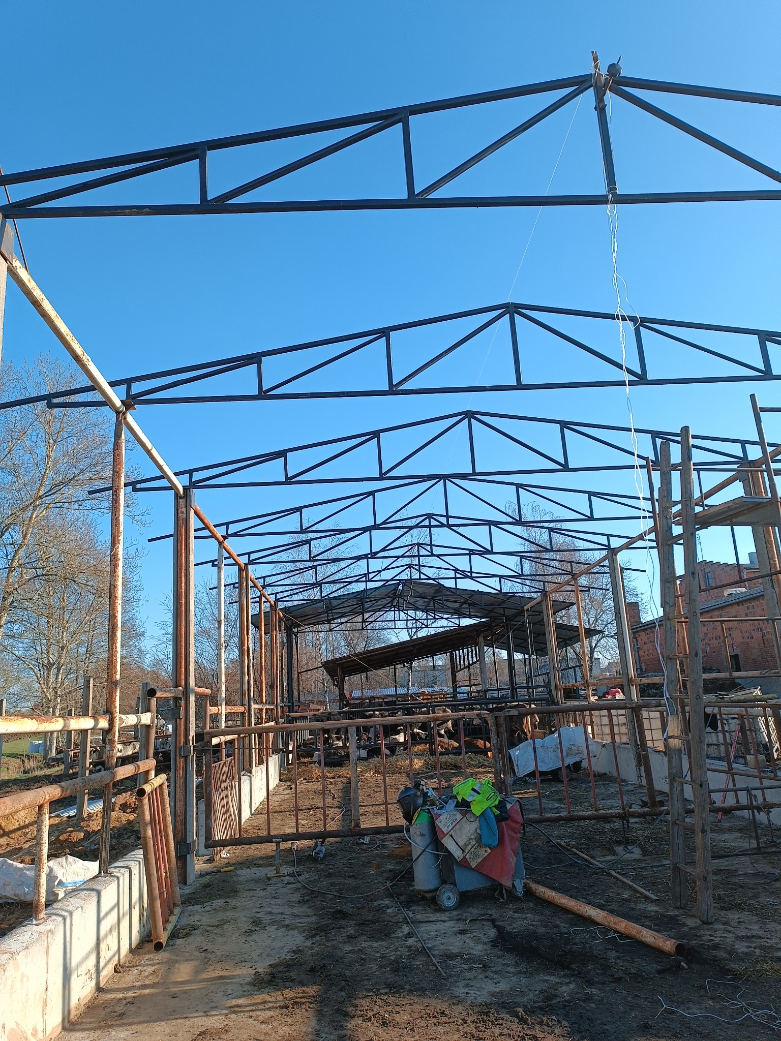 Stalowa konstrukcja szkieletowa budynku w trakcie montażu, widoczne niekompletne ściany i zadaszenie, na pierwszym planie wózek z butlami gazowymi i sprzętem spawalniczym, błękitne niebo w tle.