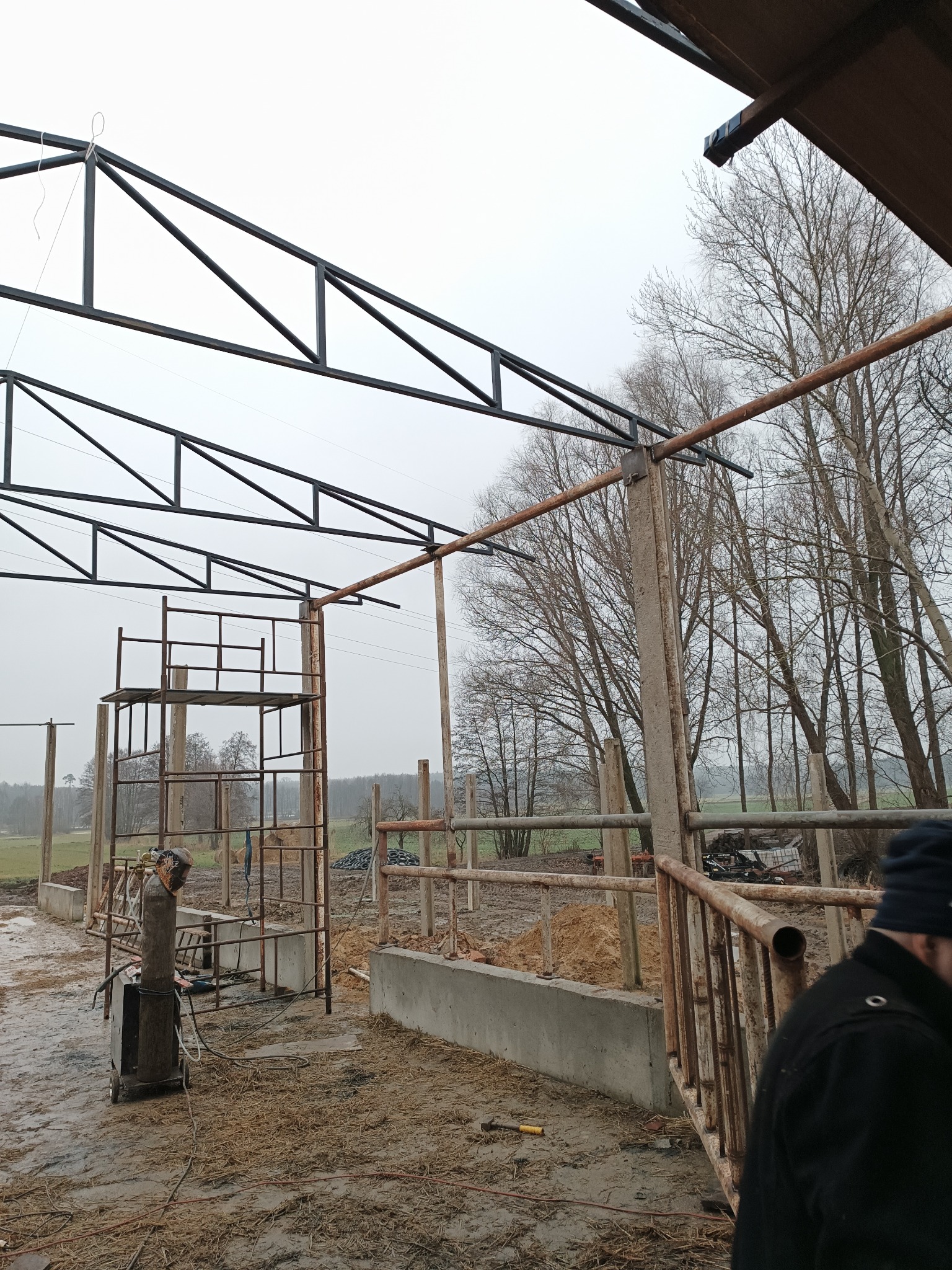 W trakcie budowy zadaszenia konstrukcji stalowej na zewnątrz, widoczny spawacz i rusztowanie, teren błotnisty.
