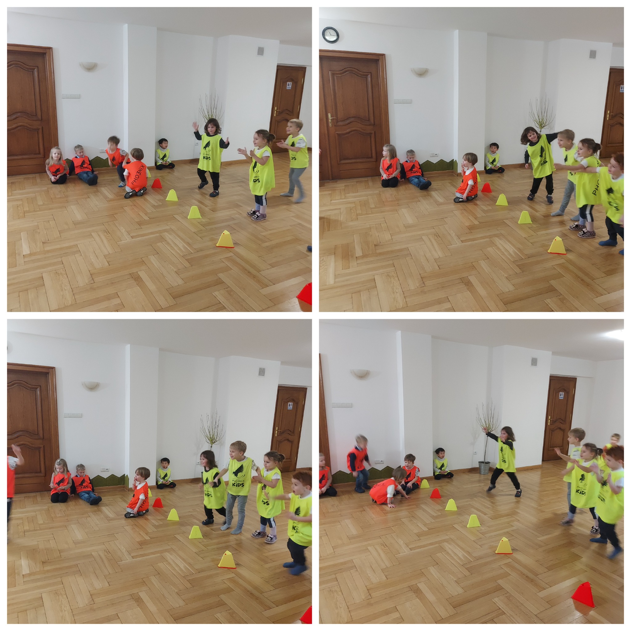 Grupa dzieci w sportowych koszulkach, część siedzi, część stoi, ćwicząc z żółtymi pachołkami na parkiecie w jasnym pomieszczeniu.