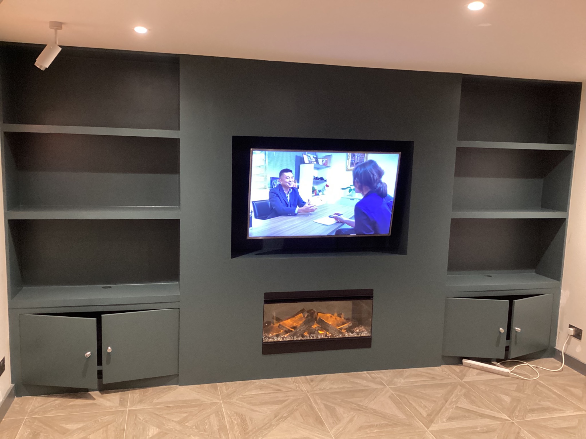 Wnęka ścienna w odcieniach szarości z półkami, szafkami z otwartymi drzwiczkami, telewizorem z włączonym ekranem i elektrycznym kominkiem poniżej