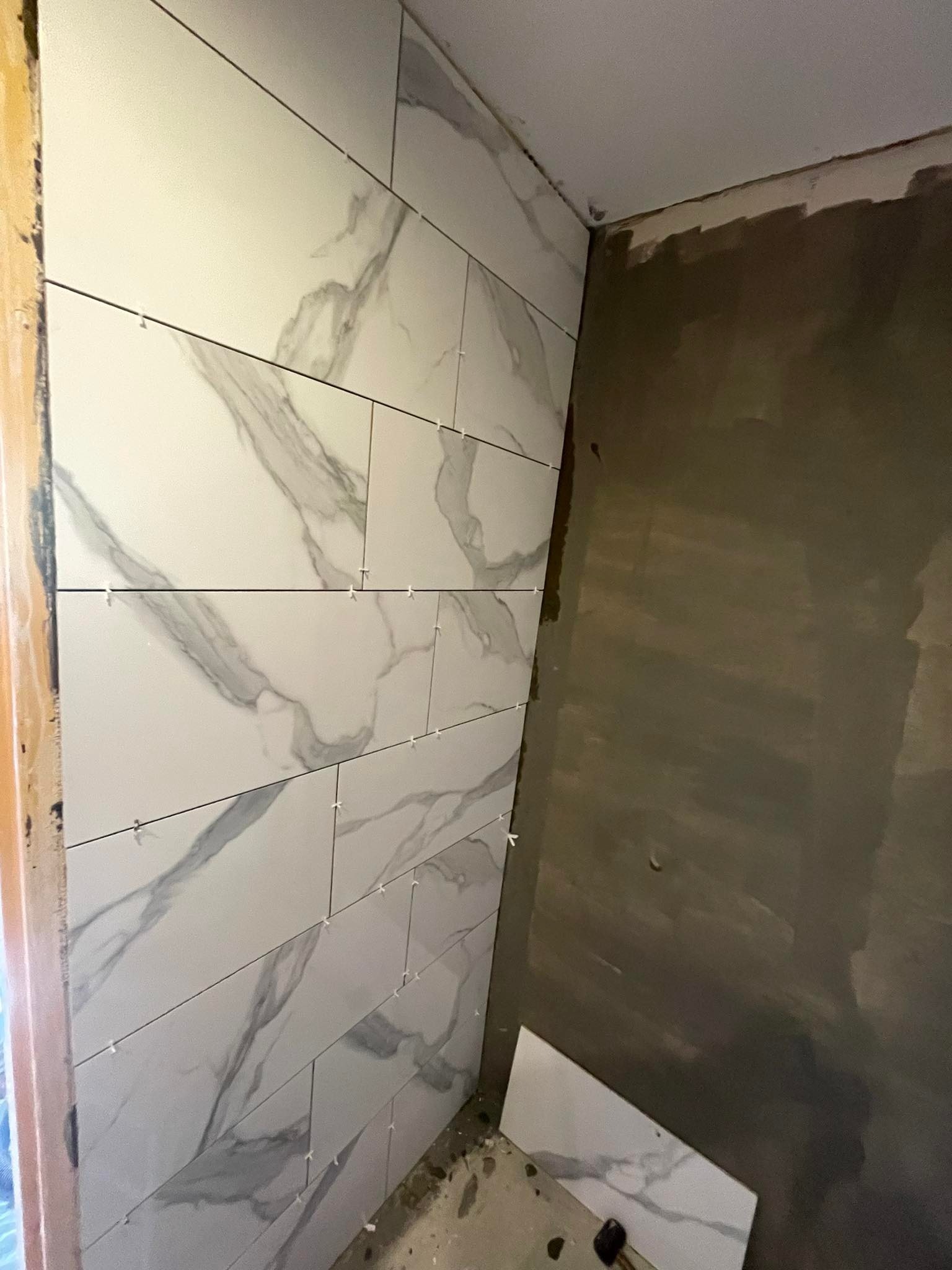 Fragment łazienki w trakcie układania białych płytek imitujących marmur, widoczne krzyżyki dystansowe i przygotowana ściana do dalszych prac.