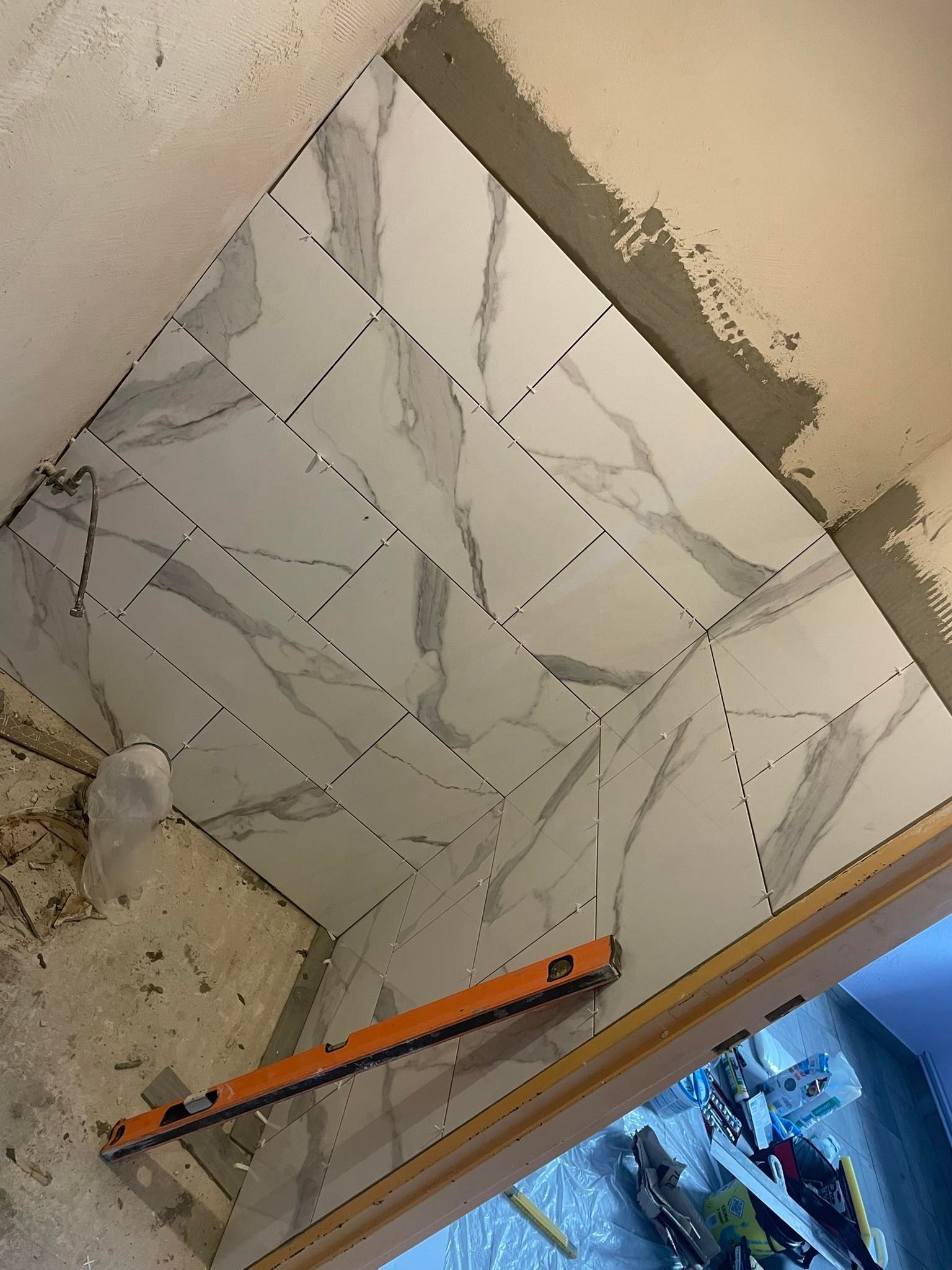 Układanie białych, marmurkowych płytek w łazience z użyciem poziomicy i krzyżyków dystansowych, widoczne ślady zaprawy na ścianie.