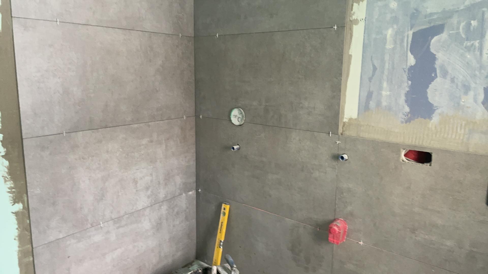 Świeżo ułożone, szare płytki w łazience z widocznymi poziomnicami laserowymi i krzyżykami dystansowymi, fragment ściany z pozostałościami po starej farbie.