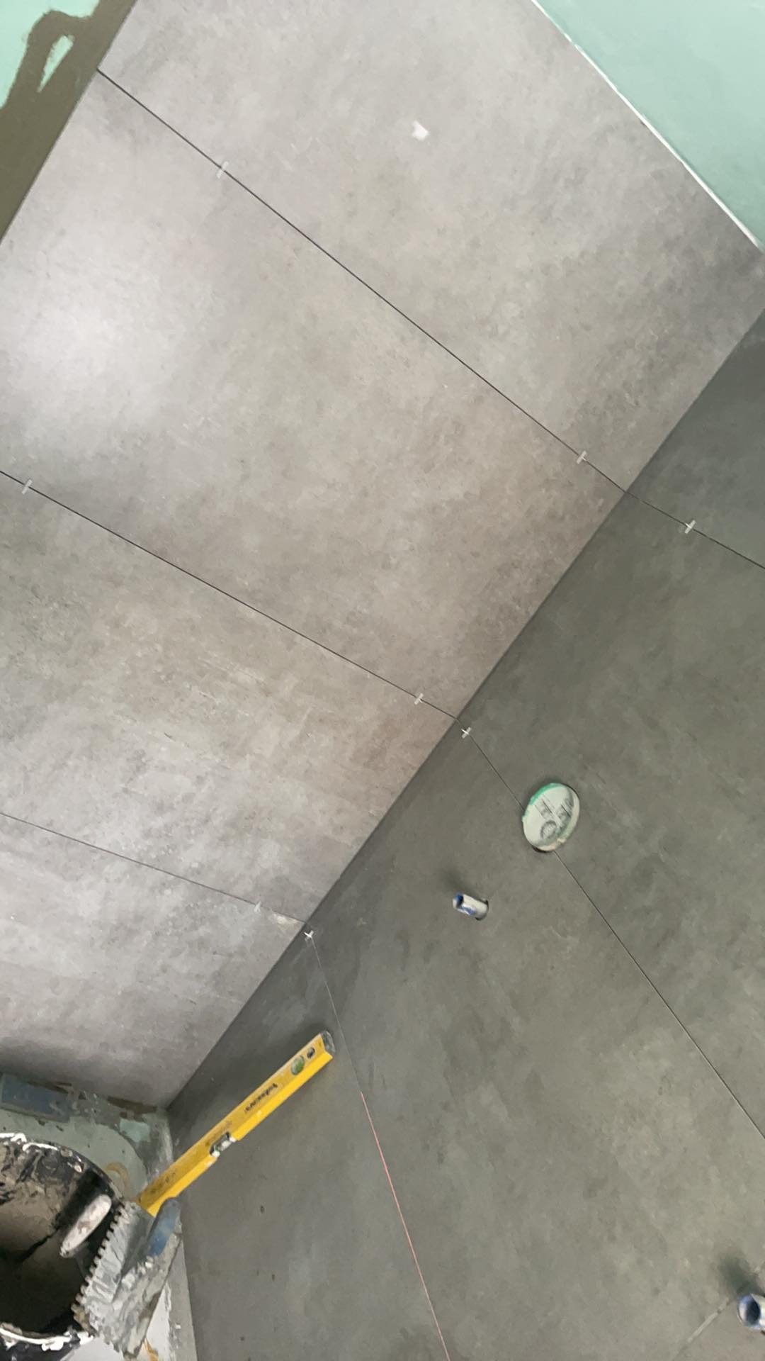 Układanie szarych płytek w łazience z użyciem poziomicy i sznurka traserskiego, widoczne krzyżyki dystansowe i puszka elektryczna.