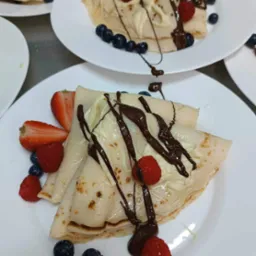 Elegancko podane naleśniki na białym talerzu, udekorowane kremem, polewą czekoladową, truskawkami, malinami i borówkami, widoczne inne talerze z naleśnikami w tle.