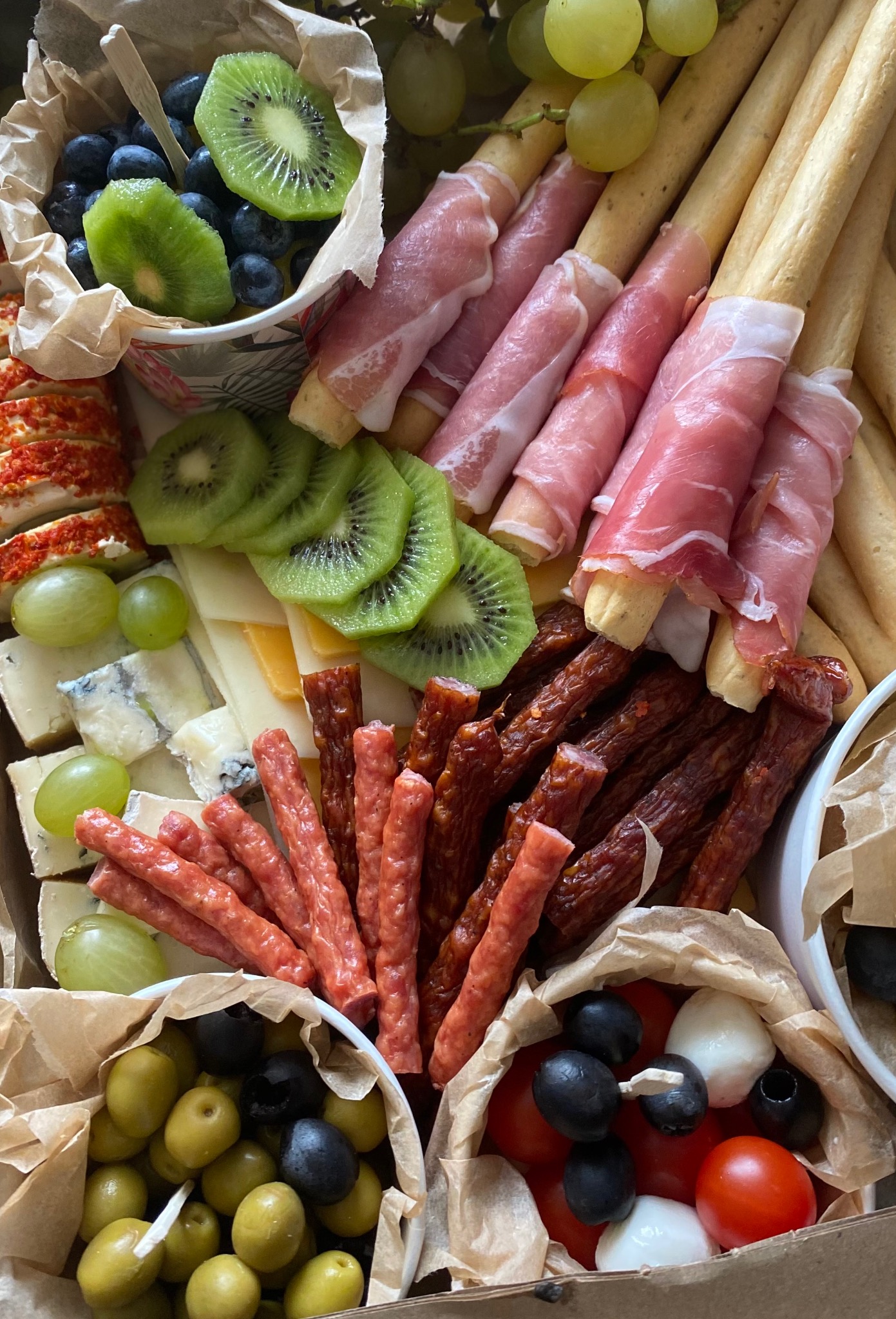 Dekoracyjna kompozycja przekąsek w kartonowym pudełku: grissini owinięte szynką parmeńską, salami, sery pleśniowe i żółte, kiwi w plasterkach, winogrona, oliwki zielone i czarne, pomidorki cherry...