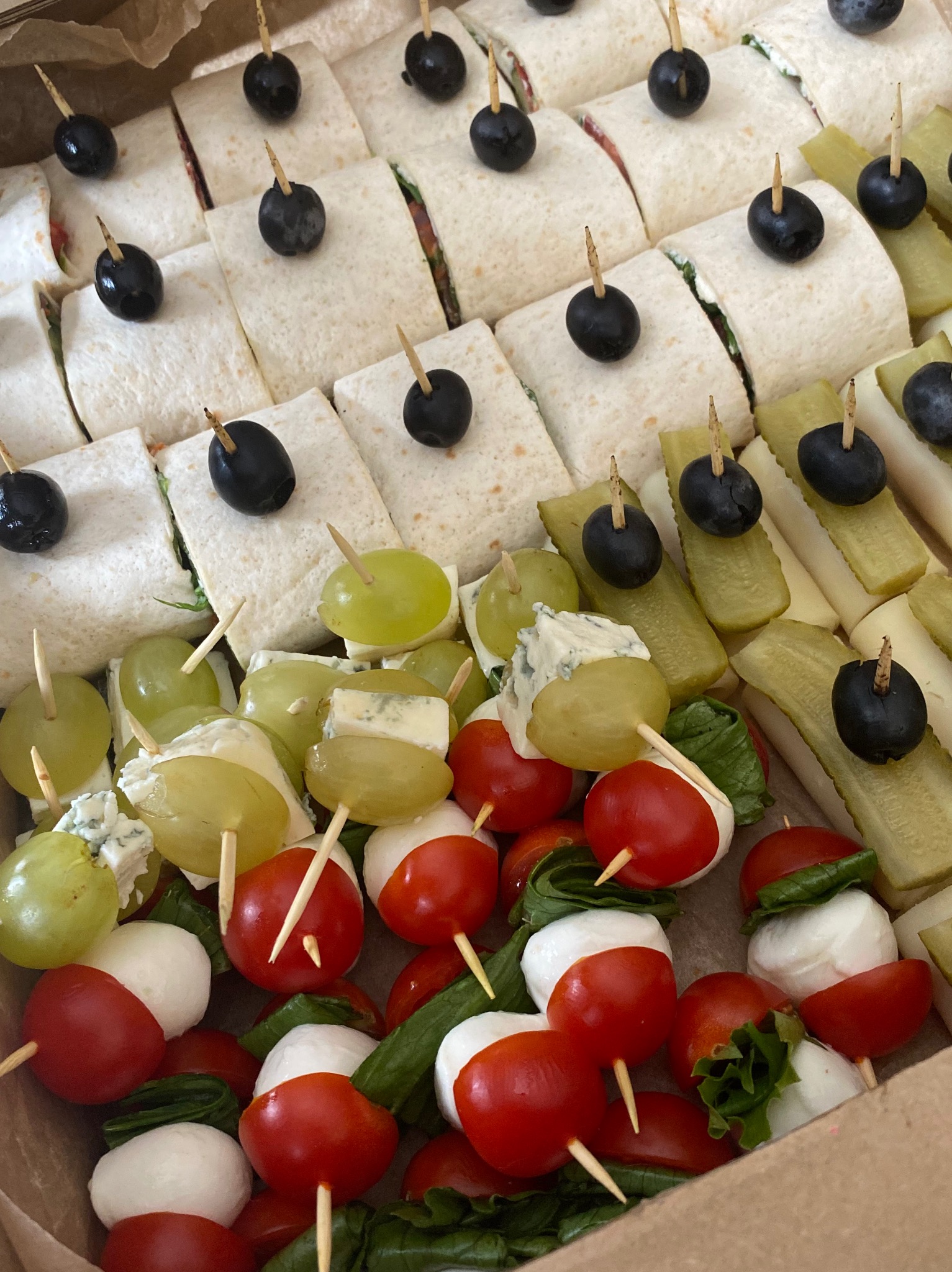 Zbliżenie na pudełko z różnorodnymi przekąskami na szpikulcach: tortille w plasterkach, koreczki z winogron, sera pleśniowego, pomidorków cherry, mini mozzarelli i bazylii, ogórków konserwowych...