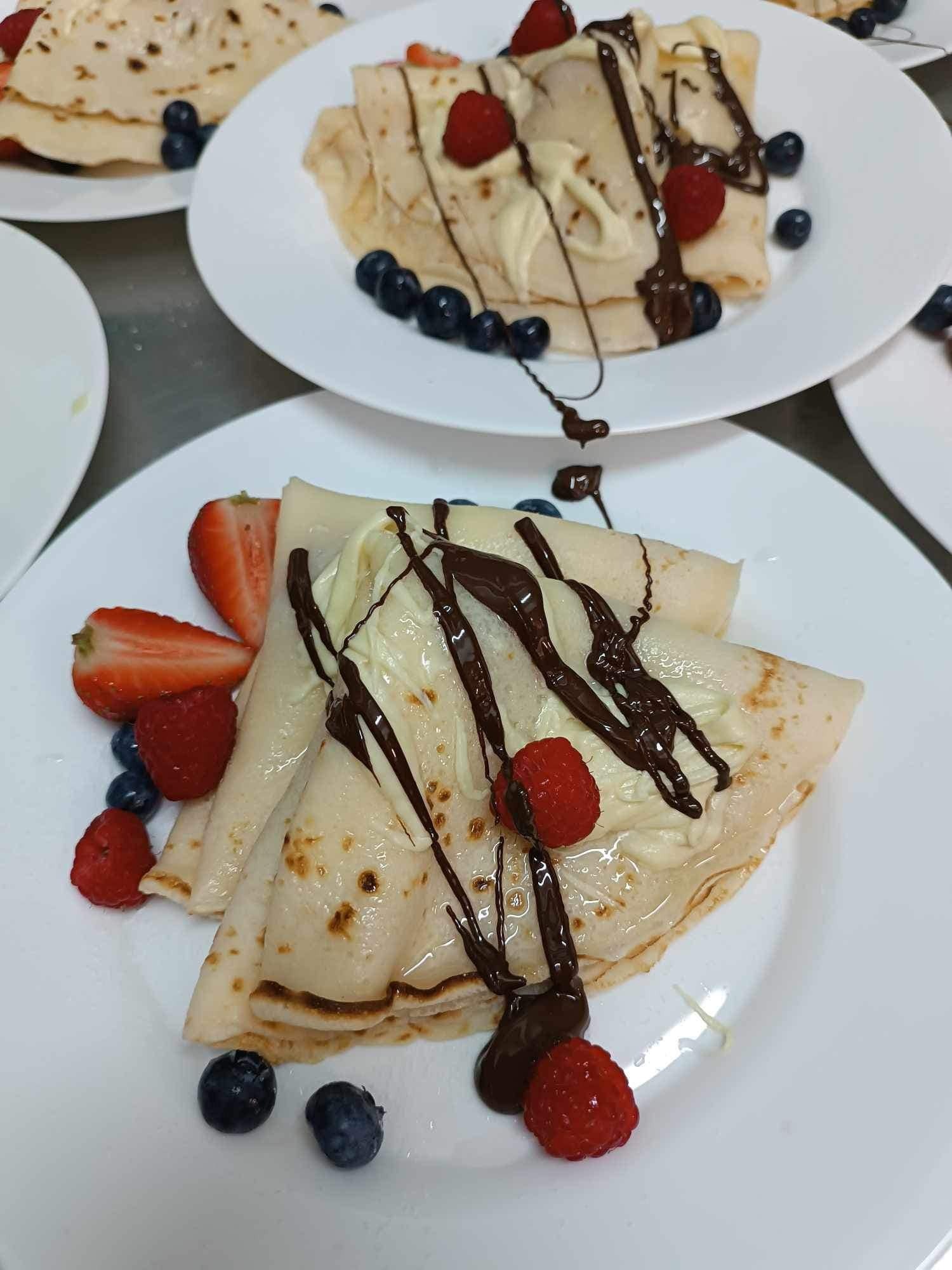 Elegancko podane naleśniki na białym talerzu, udekorowane kremem, polewą czekoladową, truskawkami, malinami i borówkami, widoczne inne talerze z naleśnikami w tle.