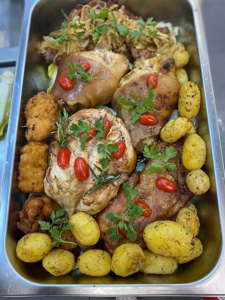 Metalowa taca z pieczonym mięsem, golonką, ziemniakami i kotletami, udekorowana pomidorkami cherry i natką pietruszki, przygotowana do serwowania.