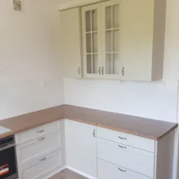 Jasnoszara zabudowa kuchenna z drewnianym blatem, widoczne szafki górne z przeszklonymi frontami i dolne z szufladami, białe płytki na ścianie, podłoga imitująca drewno.