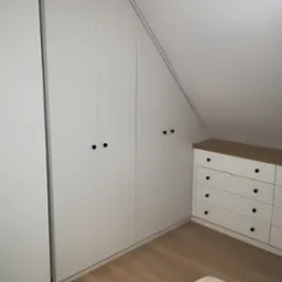 Biała szafa wnękowa z czarnymi gałkami w pokoju na poddaszu, dopasowana do skosu dachu, obok biała komoda z drewnianym blatem.