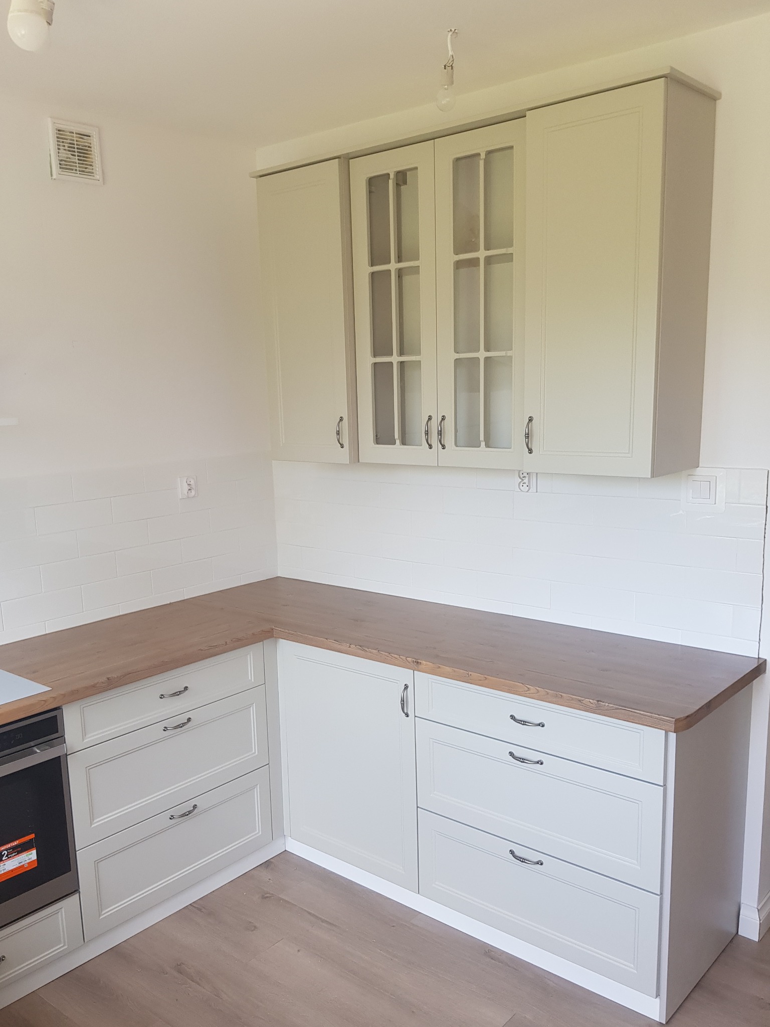 Jasnoszara zabudowa kuchenna z drewnianym blatem, widoczne szafki górne z przeszklonymi frontami i dolne z szufladami, białe płytki na ścianie, podłoga imitująca drewno.