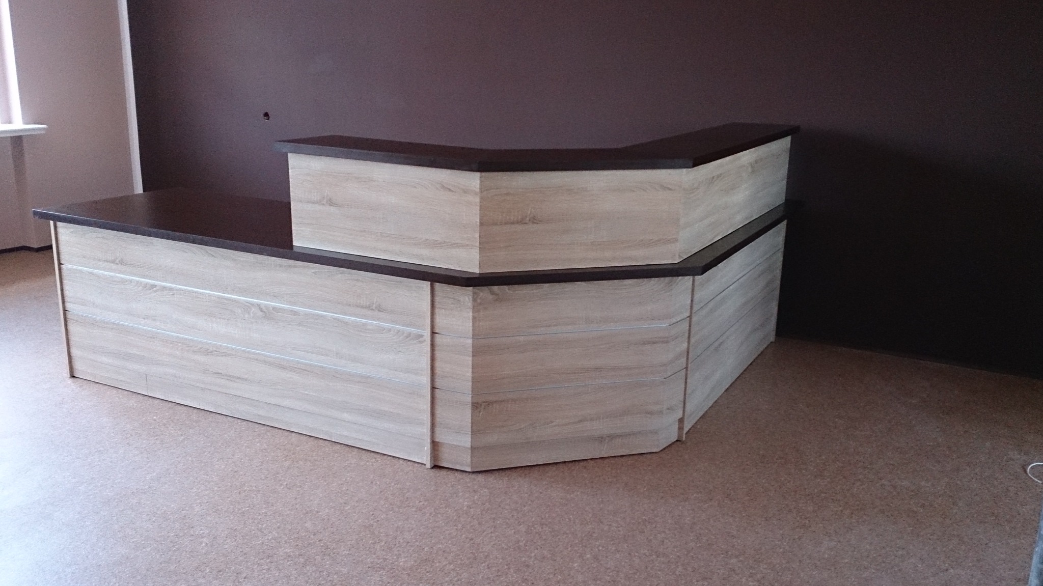 Jasna lada recepcyjna z drewnopodobnym blatem i obudową, ustawiona w rogu pomieszczenia z brązową ścianą.