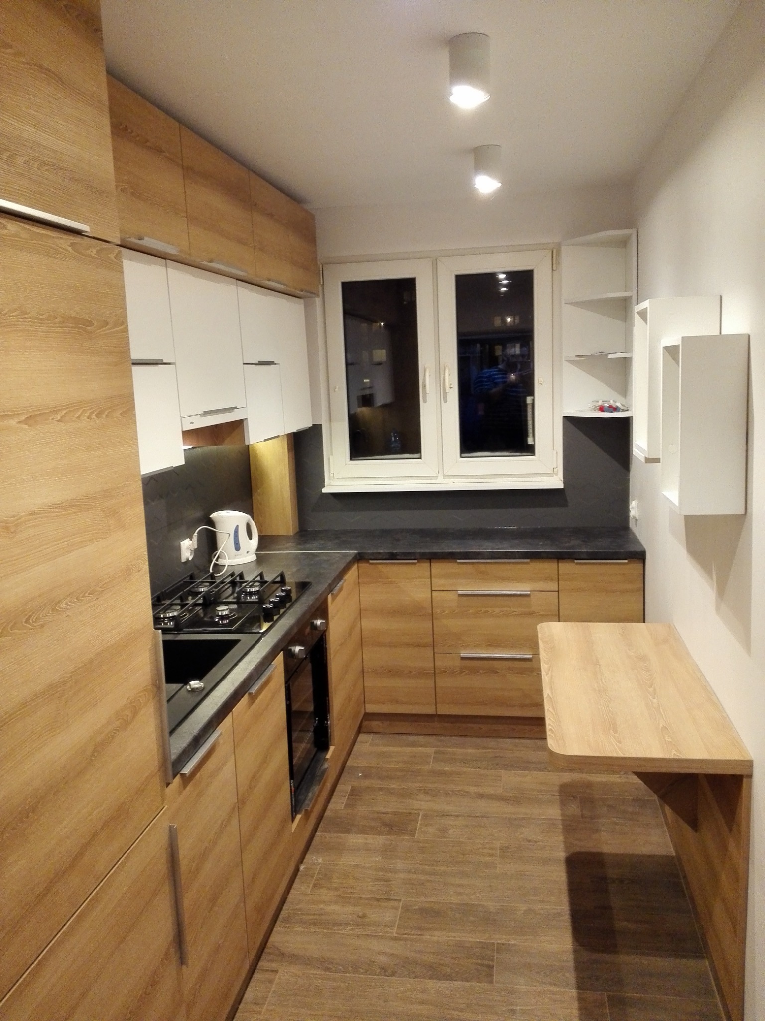 Nowoczesna kuchnia na wymiar z drewnianymi frontami i czarnym blatem, zlokalizowana w Elblągu, z dodatkowym blatem przymocowanym do ściany, oświetlona dwoma punktowymi lampami sufitowymi.