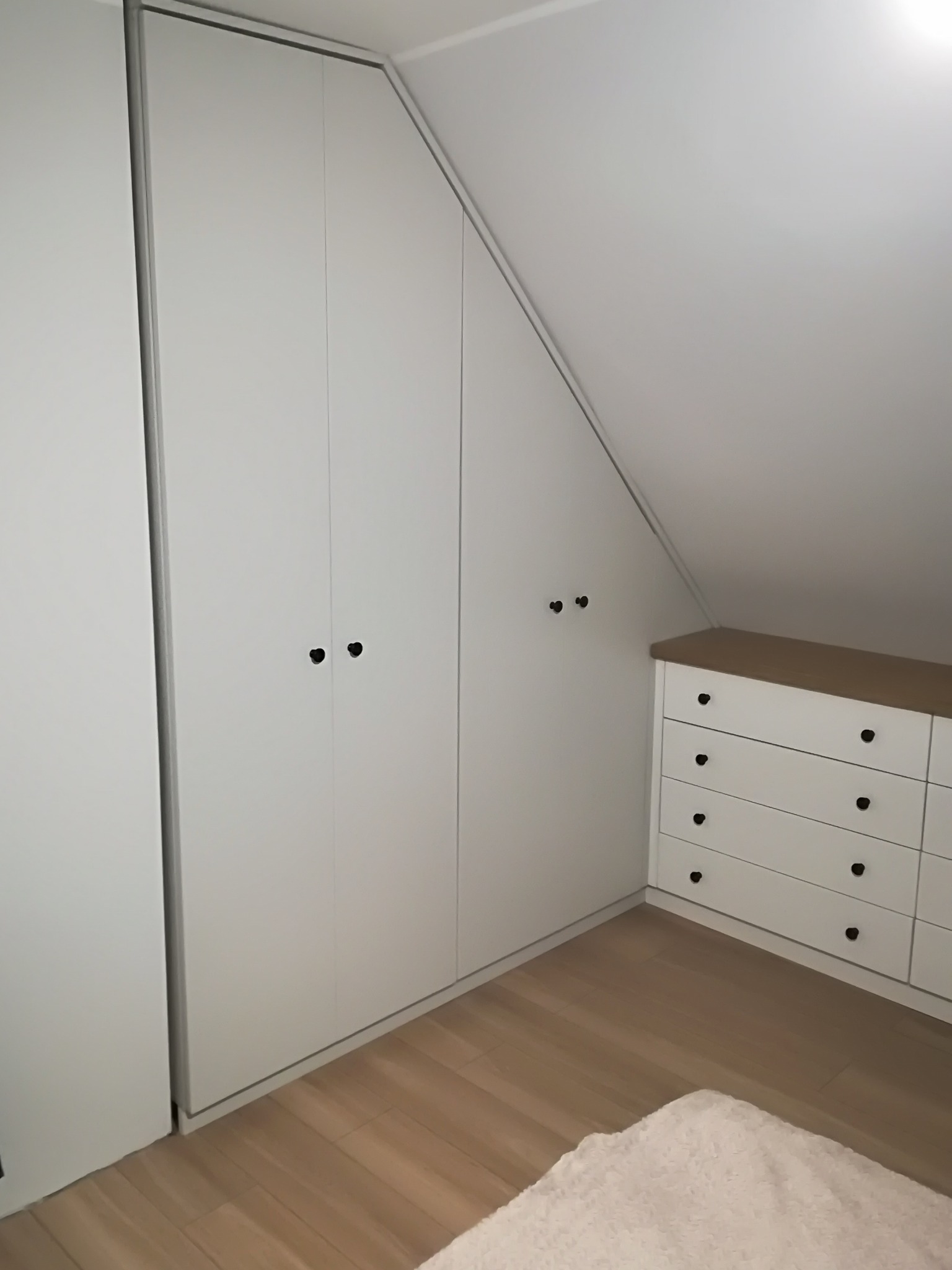 Biała szafa wnękowa z czarnymi gałkami w pokoju na poddaszu, dopasowana do skosu dachu, obok biała komoda z drewnianym blatem.