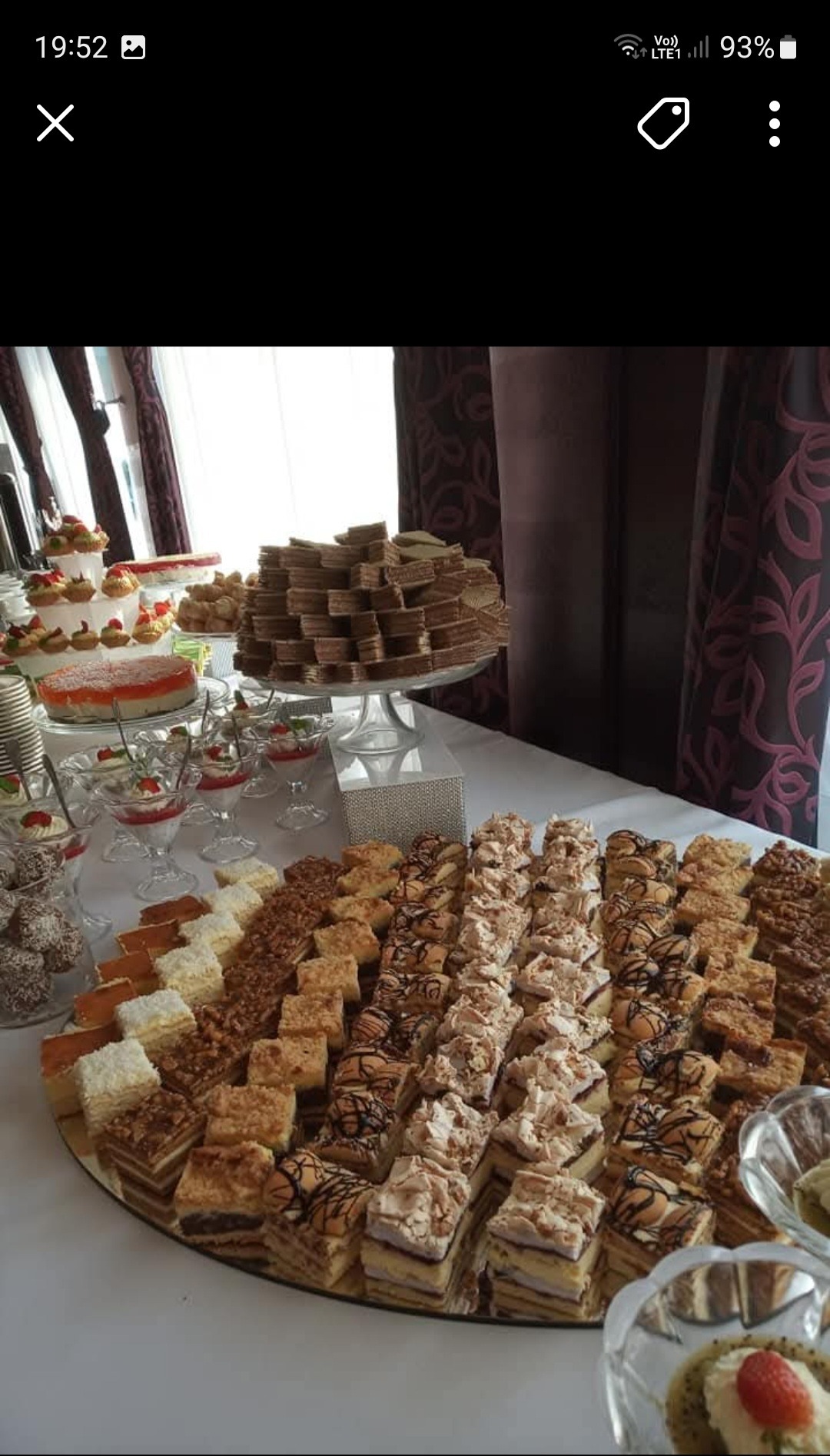 Bogato zastawiony stół z różnorodnymi ciastami, deserami i tortami, w tle zasłony i okno, sugerujące uroczystość.