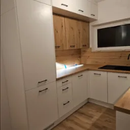 Nowoczesna kuchnia z białymi frontami i drewnianymi akcentami, czarne uchwyty, oświetlenie sufitowe w trakcie montażu, drewnopodobne panele na podłodze.