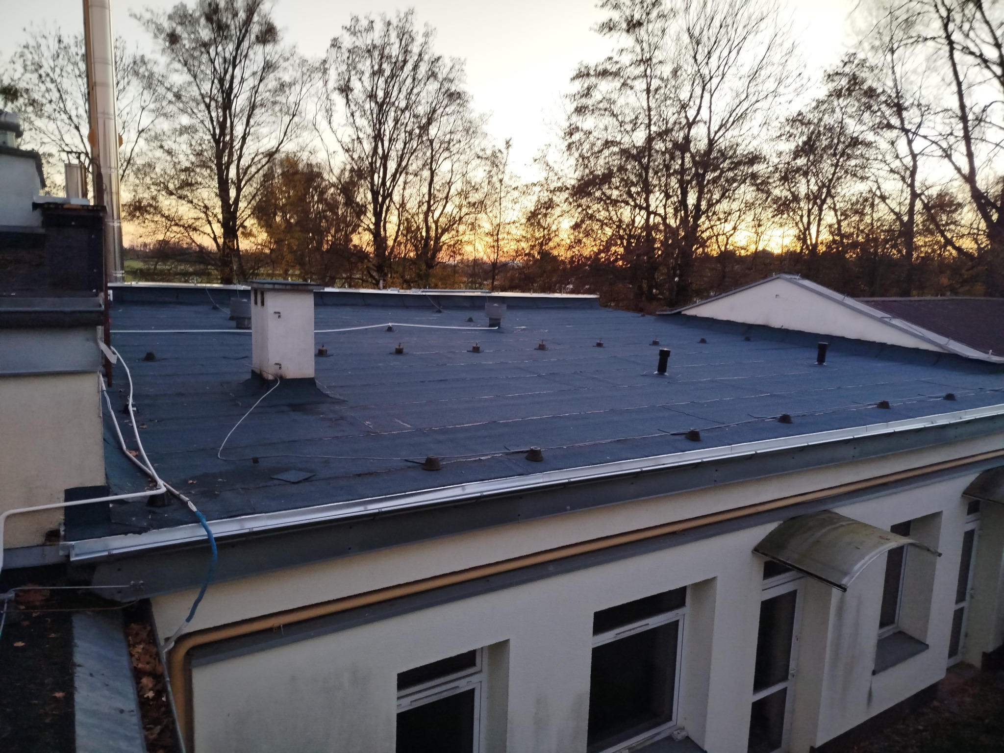 Wykończony dach pokryty papą termozgrzewalną na budynku z oknami, widok z góry, w tle drzewa i zachodzące słońce. Widoczne kominy i obróbki blacharskie.