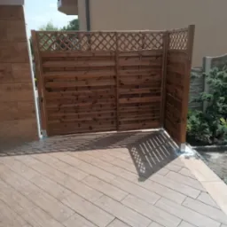 Drewniana ażurowa przesłona oddzielająca taras wyłożony płytkami imitującymi drewno od części ogrodu z roślinami.