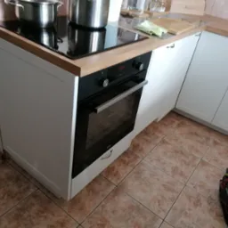 Nowoczesna kuchnia z białymi szafkami, płytą indukcyjną z dwoma garnkami oraz piekarnikiem Amica z wyświetlaczem pokazującym godzinę 13:57. Na podłodze brązowe płytki, a obok szafki torba...