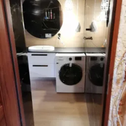 Nowoczesna łazienka z umywalką nablatową, okrągłym lustrem z czarnym tłem, dwiema pralkami Samsung i designerskimi lampami wiszącymi, widziana zza drewnianej futryny.