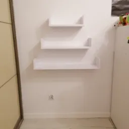 Trzy białe, minimalistyczne półki ścienne zamontowane na białej ścianie nad gniazdkiem elektrycznym, obok beżowej szafy przesuwnej i białej szafki.