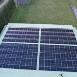 Widok z góry na dużą instalację fotowoltaiczną na dachu budynku przemysłowego, z widocznym fragmentem trawnika i drzew oraz samochodem na parkingu.