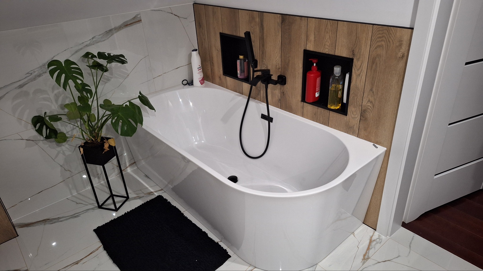 Biała wanna wolnostojąca z czarną armaturą, rośliną Monstera w czarnej donicy na tle płytek imitujących marmur i drewnianej ściany z wnękami na kosmetyki.