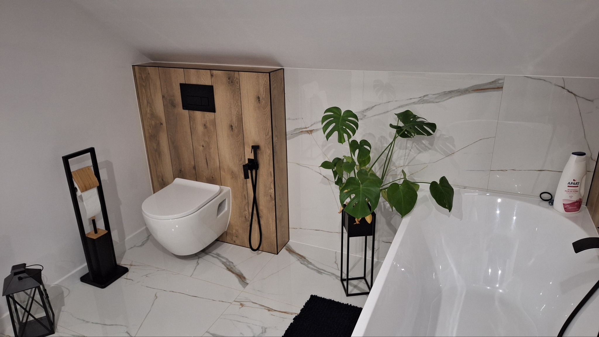 Nowoczesna łazienka na poddaszu z białymi płytkami imitującymi marmur, wiszącą toaletą z drewnianą zabudową, czarną armaturą i dekoracyjną rośliną Monstera.