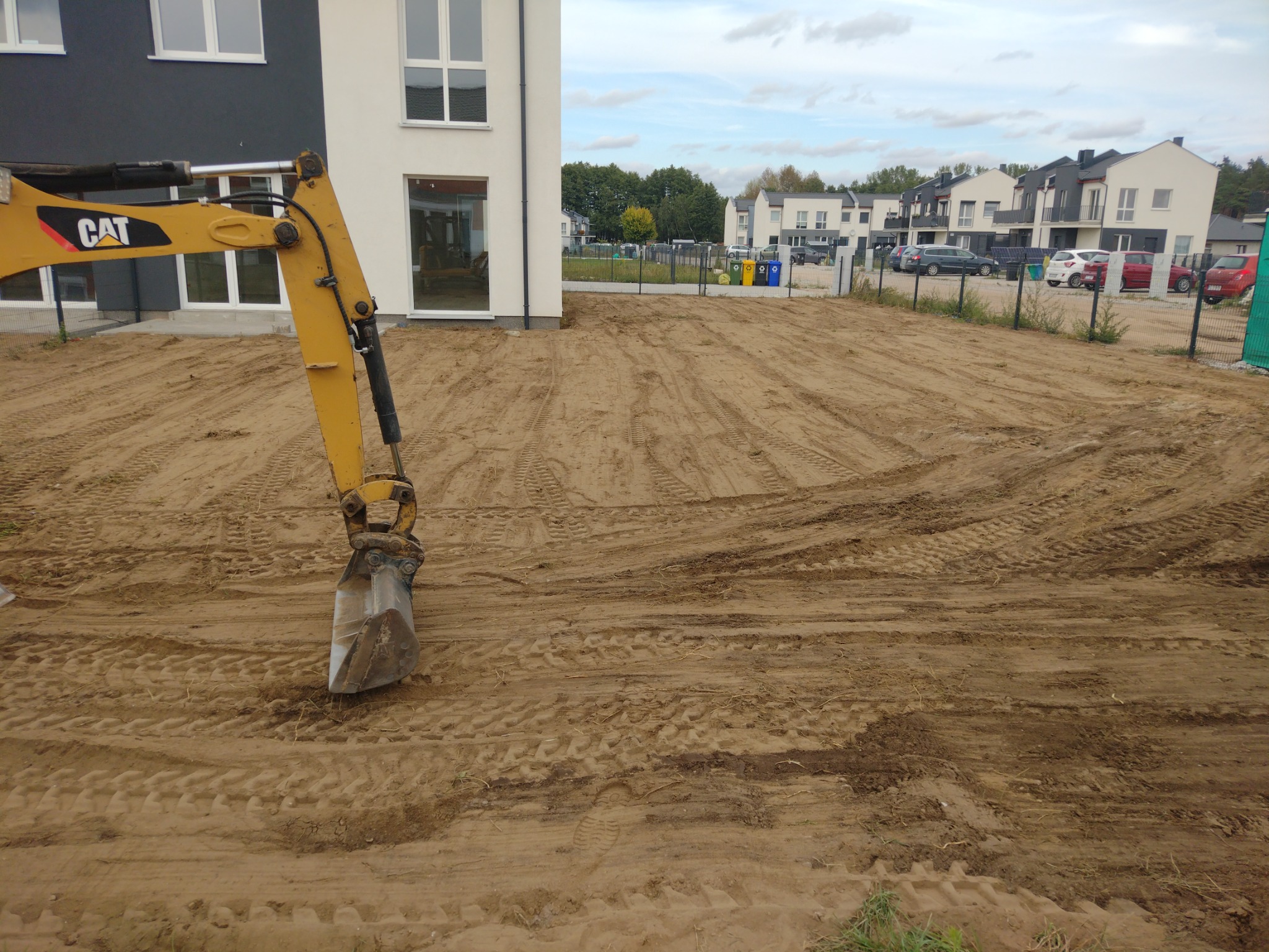 Żółta koparka CAT z łyżką na placu budowy, wyrównany teren z widocznymi śladami opon, w tle nowe domy jednorodzinne i pochmurne niebo.
