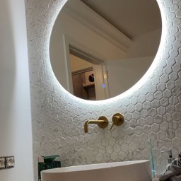 PZ PROJECT - Nowoczesna łazienka w trakcie wykańczania: okrągłe lustro z podświetleniem LED, biała umywalka nablatowa, złota armatura, ściana wyłożona mozaiką heksagonalną, widoczne niedokończone prace...