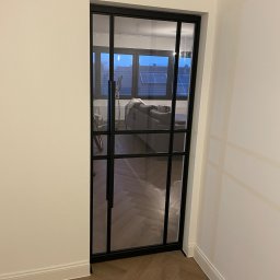 PZ PROJECT - Nowoczesne, przeszklone drzwi wewnętrzne z czarną ramą, widok na salon z parkietem ułożonym w jodełkę, rowerem i szarą sofą.