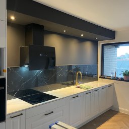 PZ PROJECT - Nowoczesna kuchnia z białymi szafkami, czarnym panelem nad blatem imitującym marmur, złotą baterią i czarnym okapem; aluminiowa drabina w rogu wskazuje na trwające prace wykończeniowe.