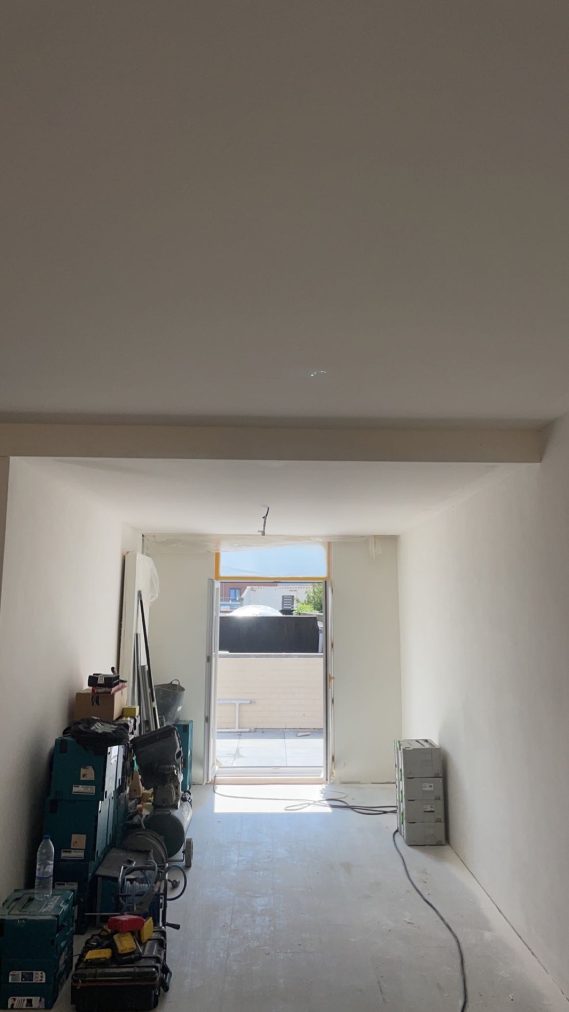 Wnętrze w trakcie prac wykończeniowych: świeżo zagruntowane ściany, sprzęt budowlany zgromadzony w rogu pomieszczenia, widoczne kable i skrzynki narzędziowe.