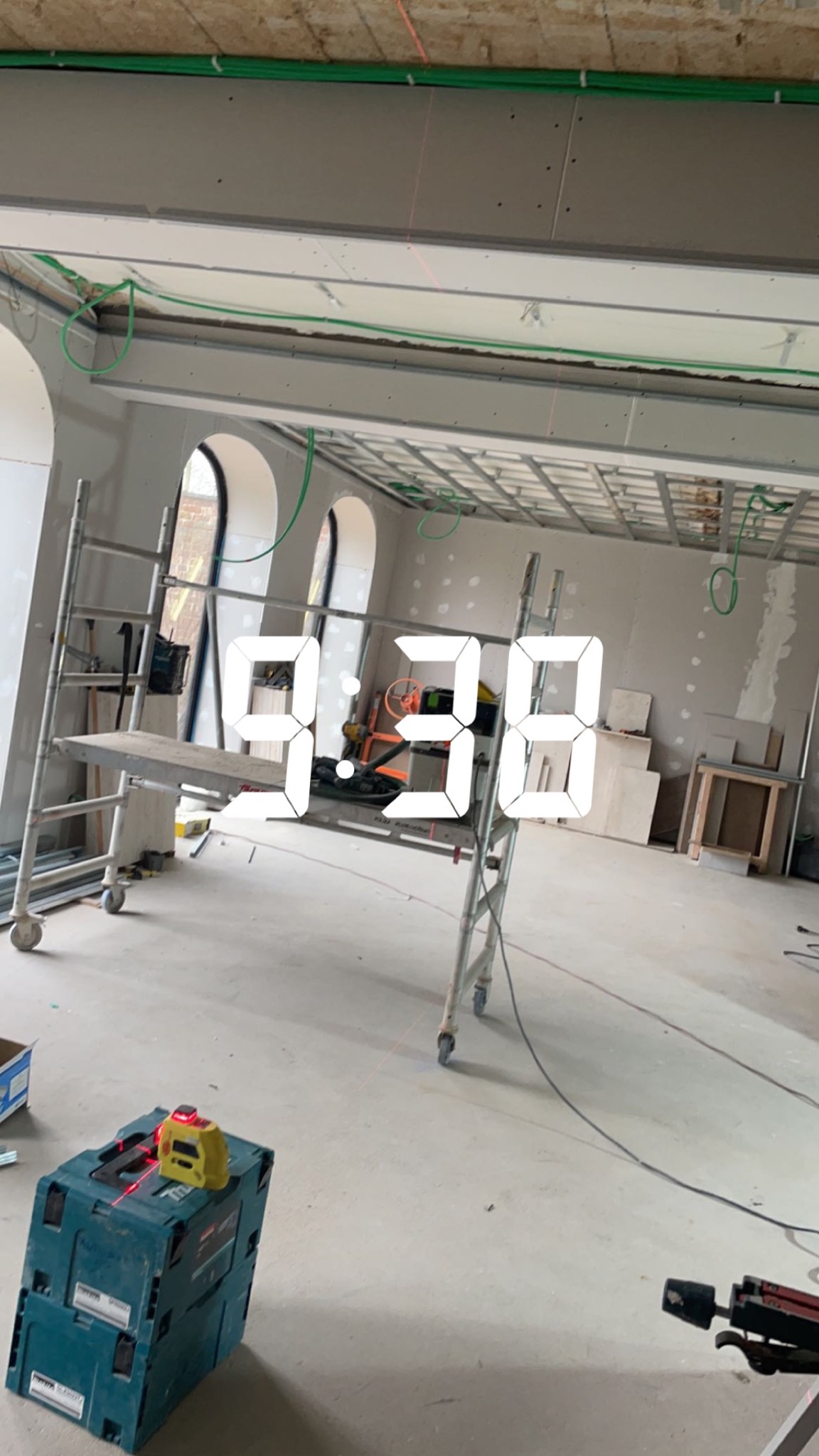 Wnętrze w trakcie prac wykończeniowych: widoczne rusztowanie, instalacje pod sufitem, narzędzia i płyty kartonowo-gipsowe przygotowane do montażu, laser krzyżowy na skrzynce narzędziowej.