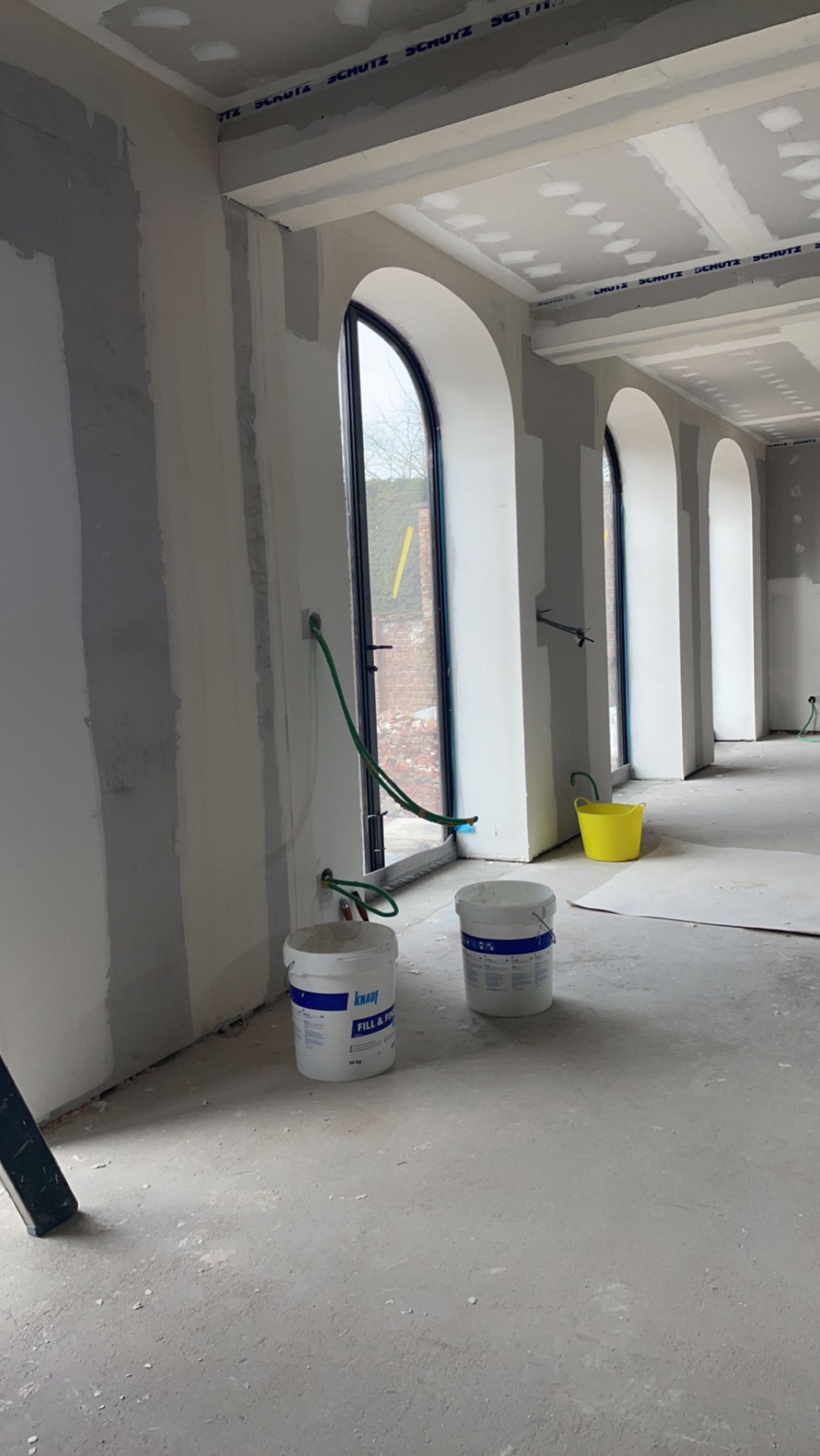 Wnętrze w trakcie prac wykończeniowych: ściany w szarych pasach po nałożeniu gładzi, sufit z płyt kartonowo-gipsowych, wysokie okna łukowe, dwa wiadra z masą szpachlową Knauf Fill & Finish...