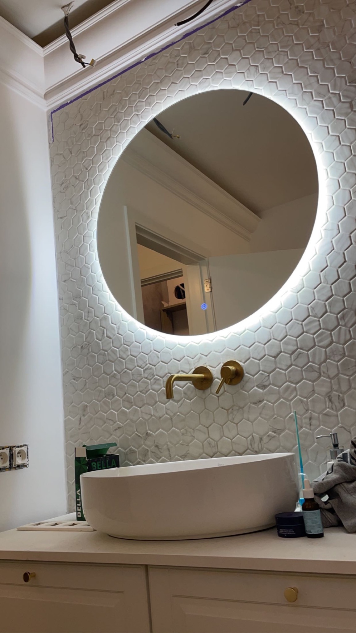 Nowoczesna łazienka w trakcie wykańczania: okrągłe lustro z podświetleniem LED, biała umywalka nablatowa, złota armatura, ściana wyłożona mozaiką heksagonalną, widoczne niedokończone prace...