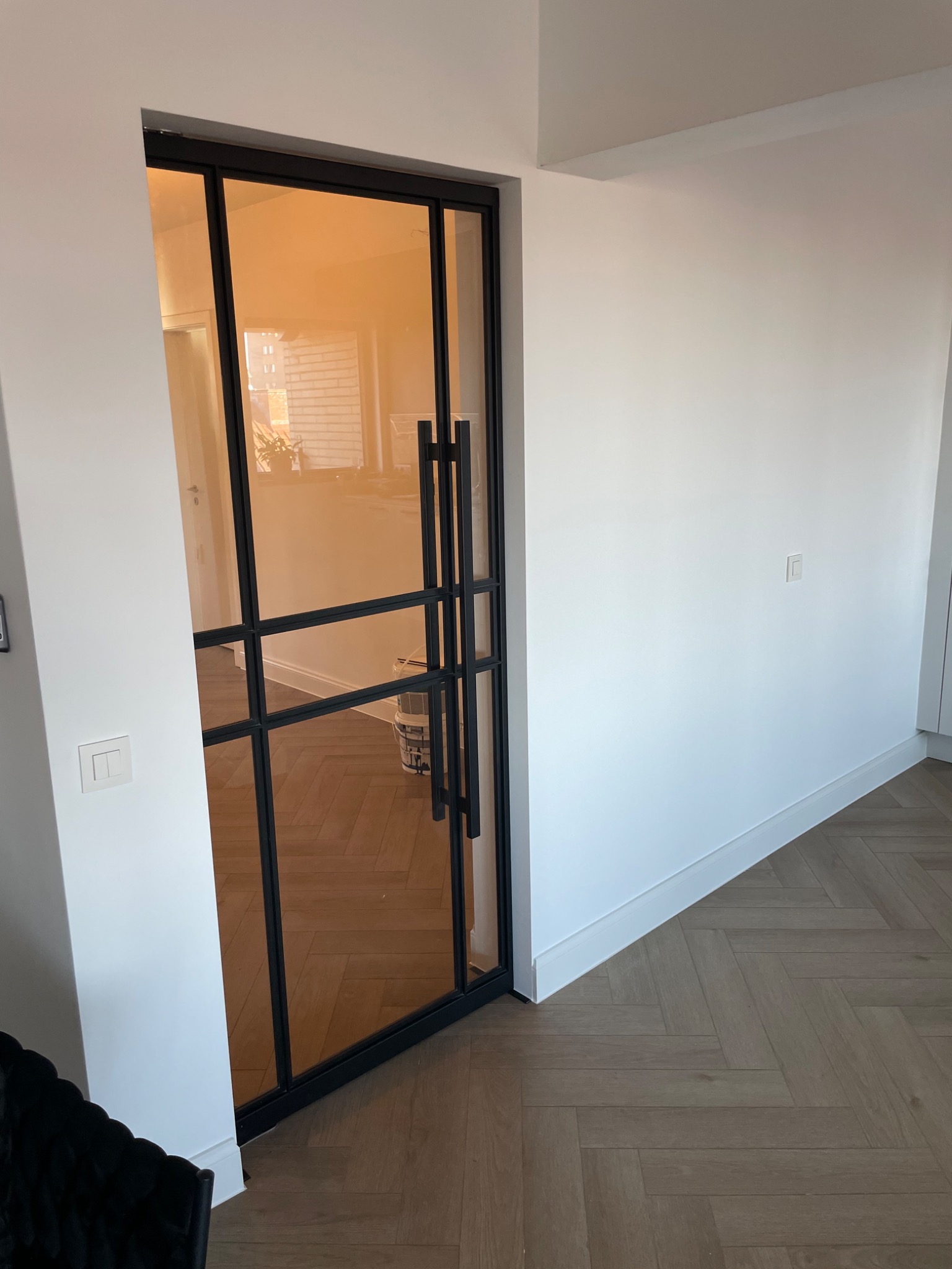 Nowoczesne drzwi wewnętrzne z czarną ramą i oranżowym szkłem, widok z boku na jasne wnętrze z parkietem w jodełkę.