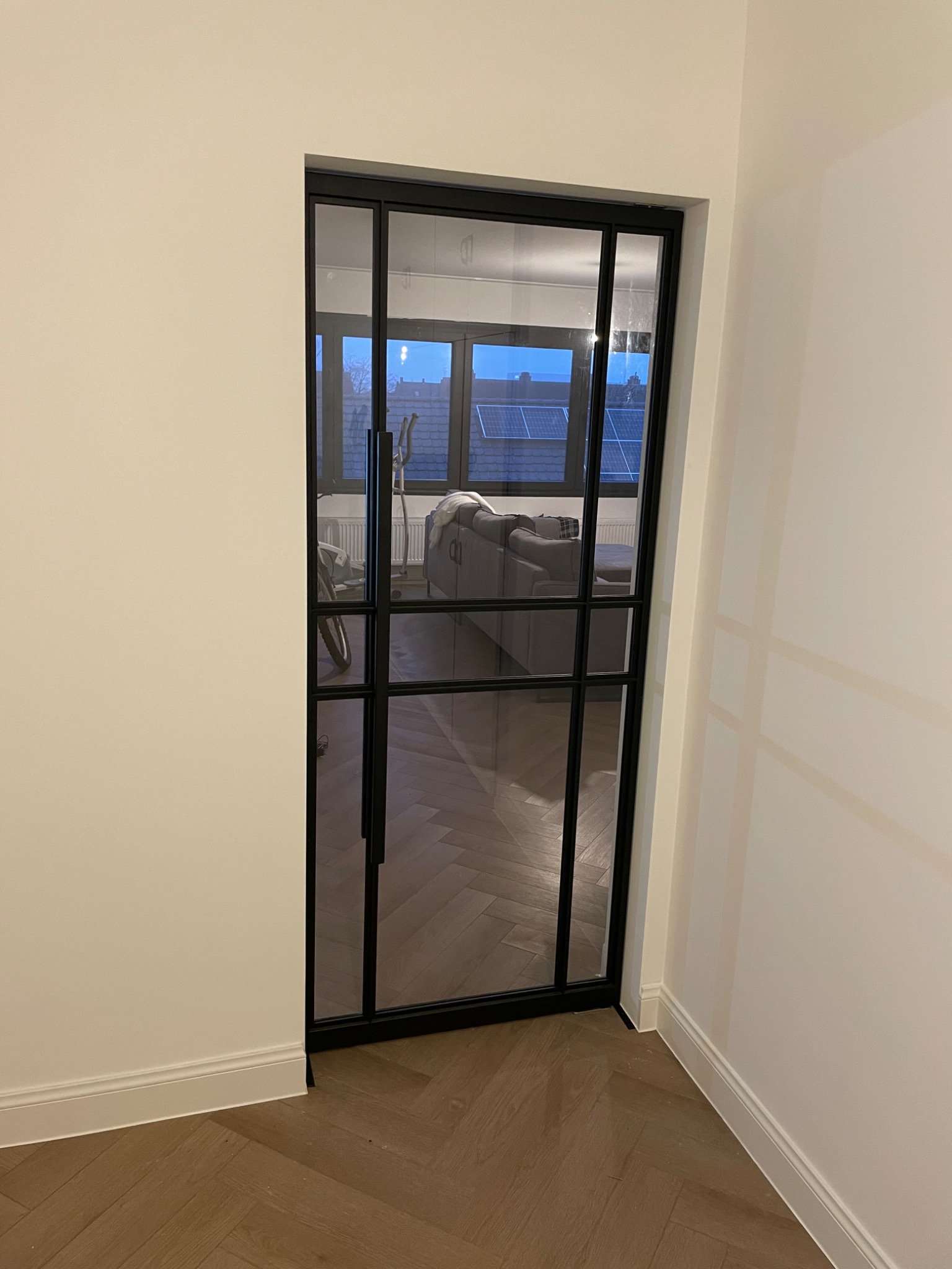 Nowoczesne, przeszklone drzwi wewnętrzne z czarną ramą, widok na salon z parkietem ułożonym w jodełkę, rowerem i szarą sofą.