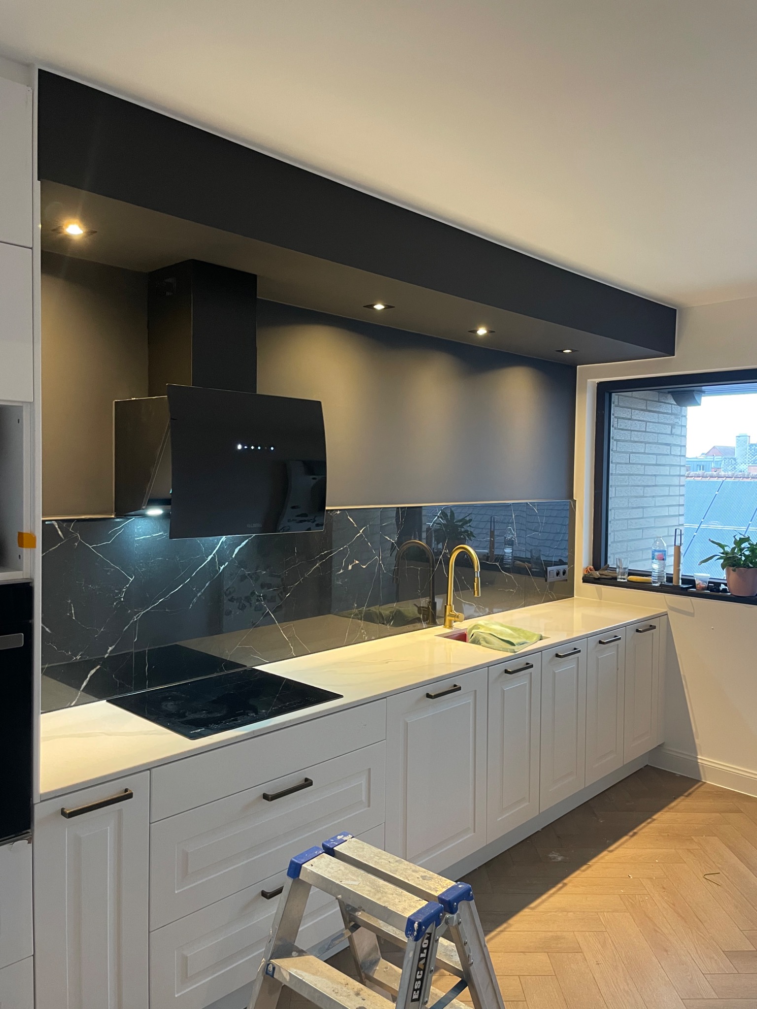 Nowoczesna kuchnia z białymi szafkami, czarnym panelem nad blatem imitującym marmur, złotą baterią i czarnym okapem; aluminiowa drabina w rogu wskazuje na trwające prace wykończeniowe.