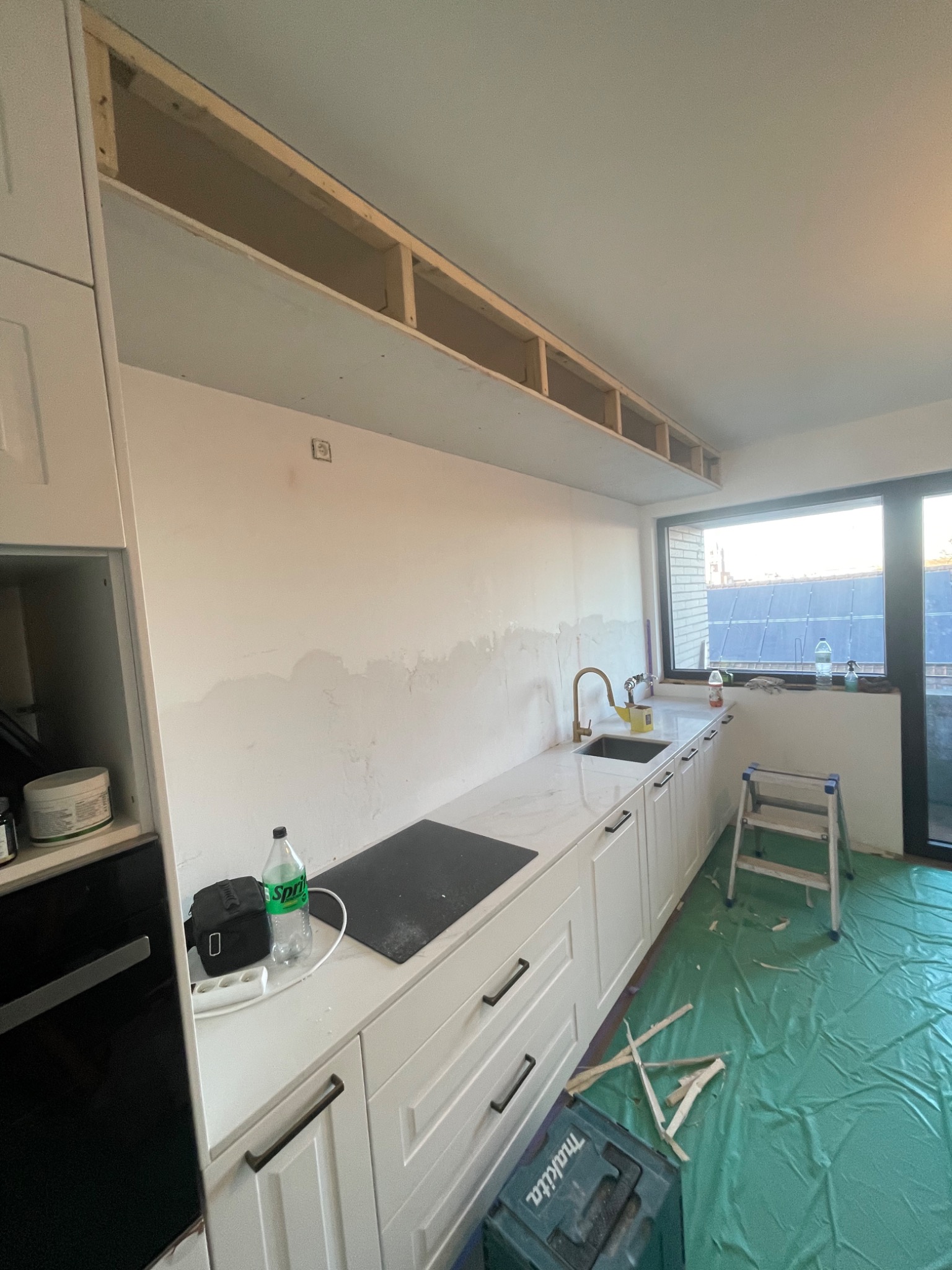 Wnętrze kuchni w trakcie remontu: widoczne białe szafki kuchenne, blat z imitacją marmuru, przygotowana ściana do gładzi, drewniana konstrukcja pod sufitem, zabezpieczona podłoga folią, drabina...